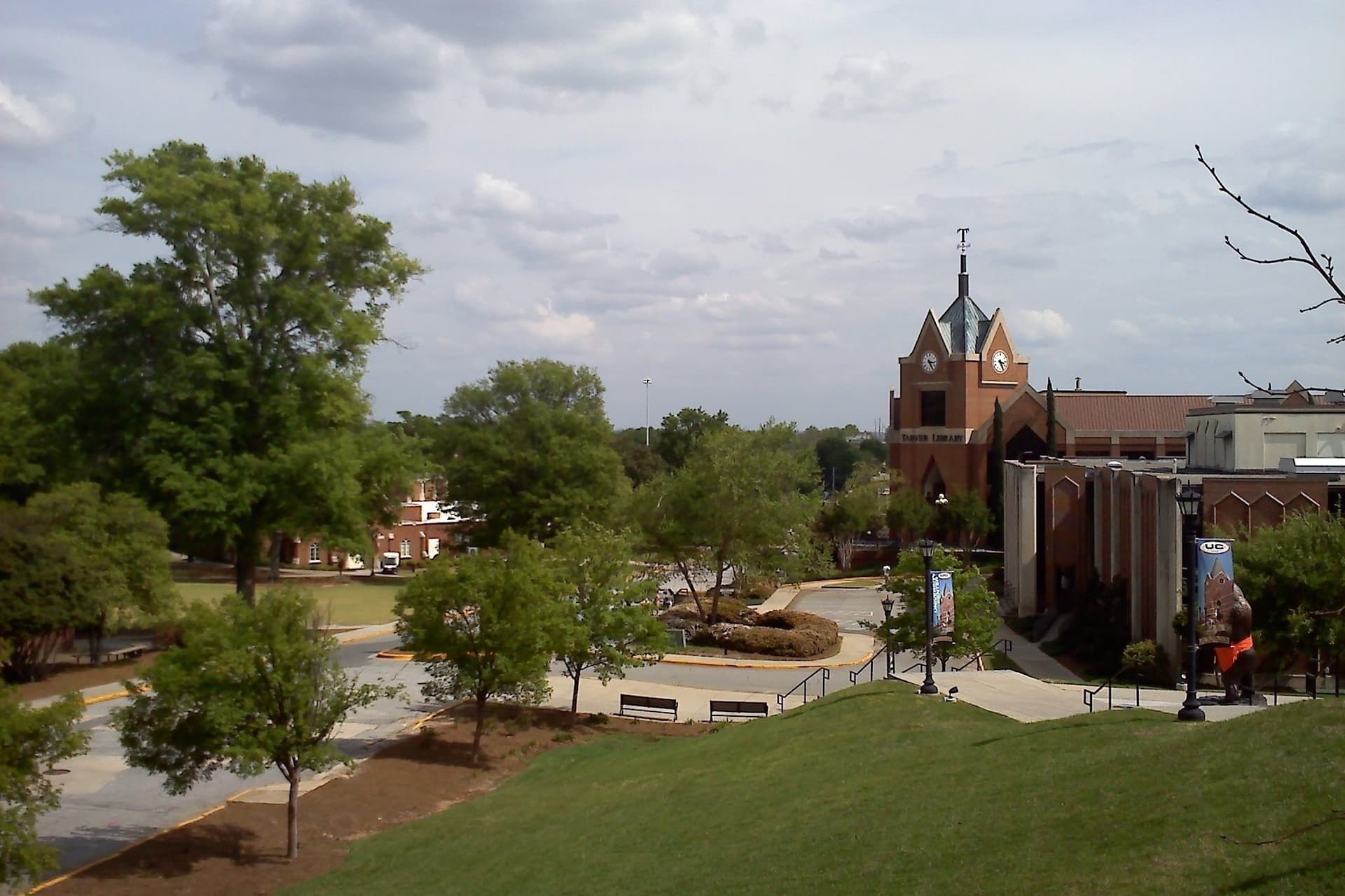 A grassy hillside and tree-lined walkways leading down to a red brick building with a clock tower and steeple under a partly cloudy sky