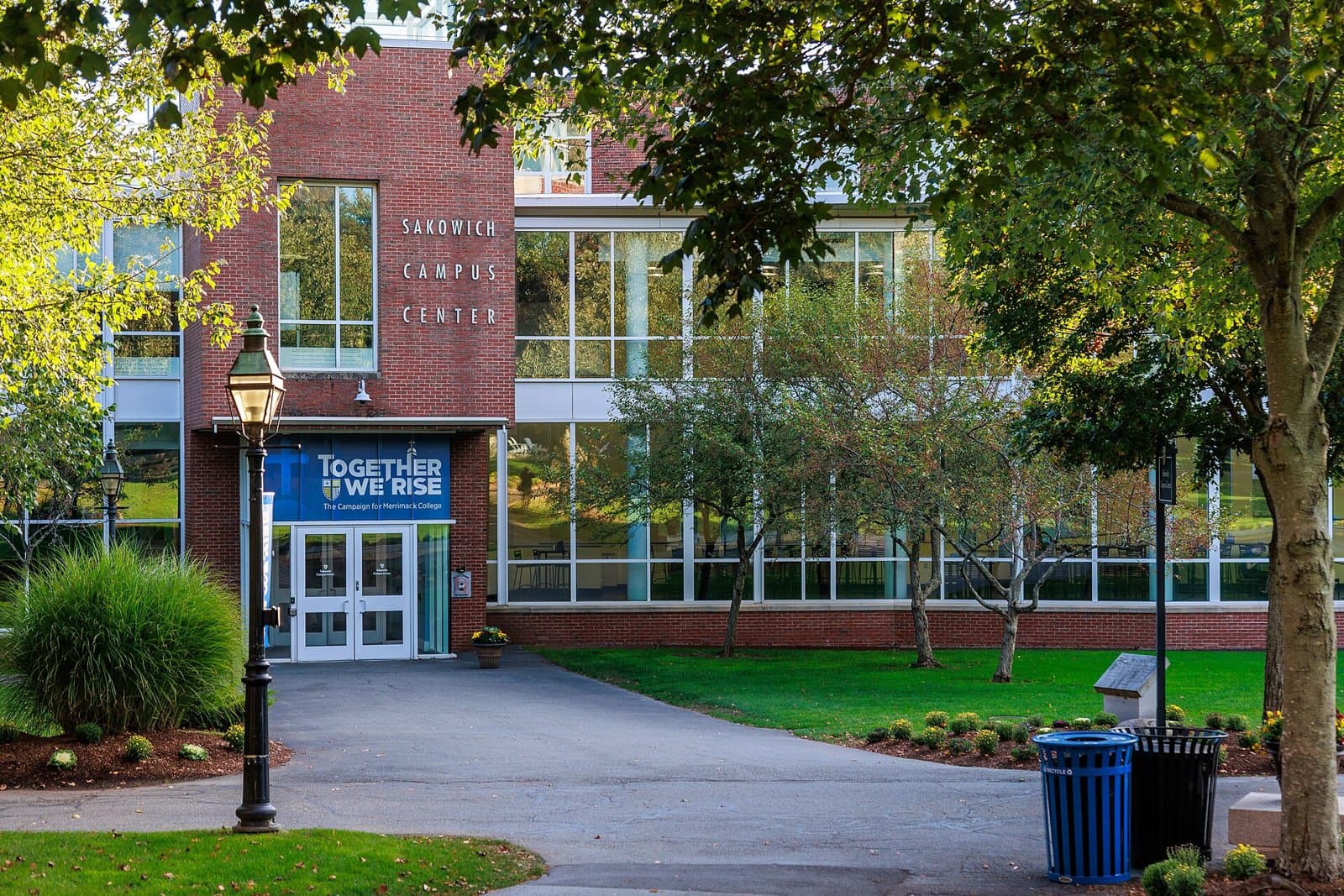 The brick and glass Sakowich Campus Center at Merrimack College framed by trees, with a lamppost and walkway leading to its entrance