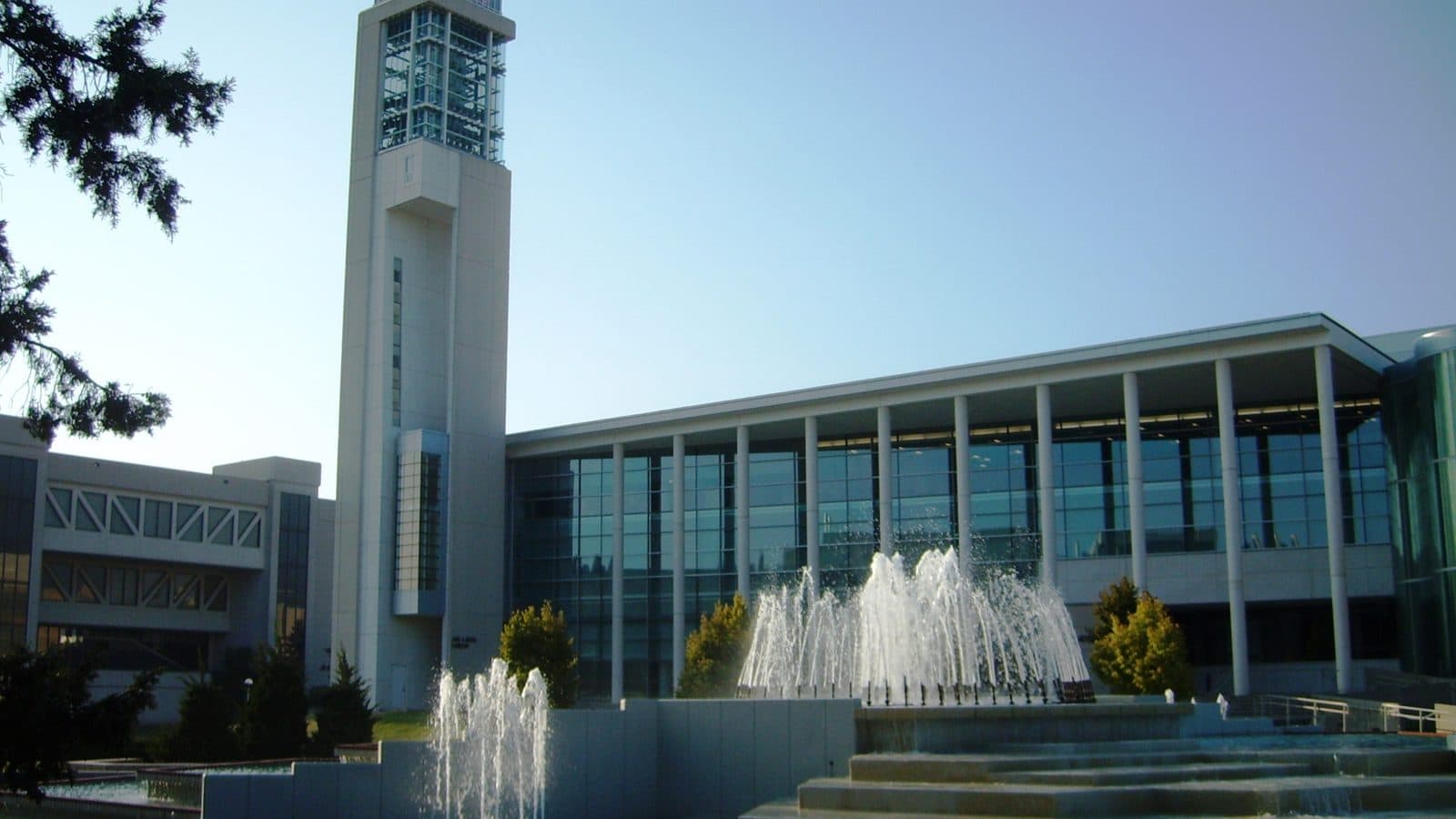 A tall white carillon tower with a glass top beside a long glass-fronted library with white columns, with tiered fountains spraying in the foreground under a clear blue sky