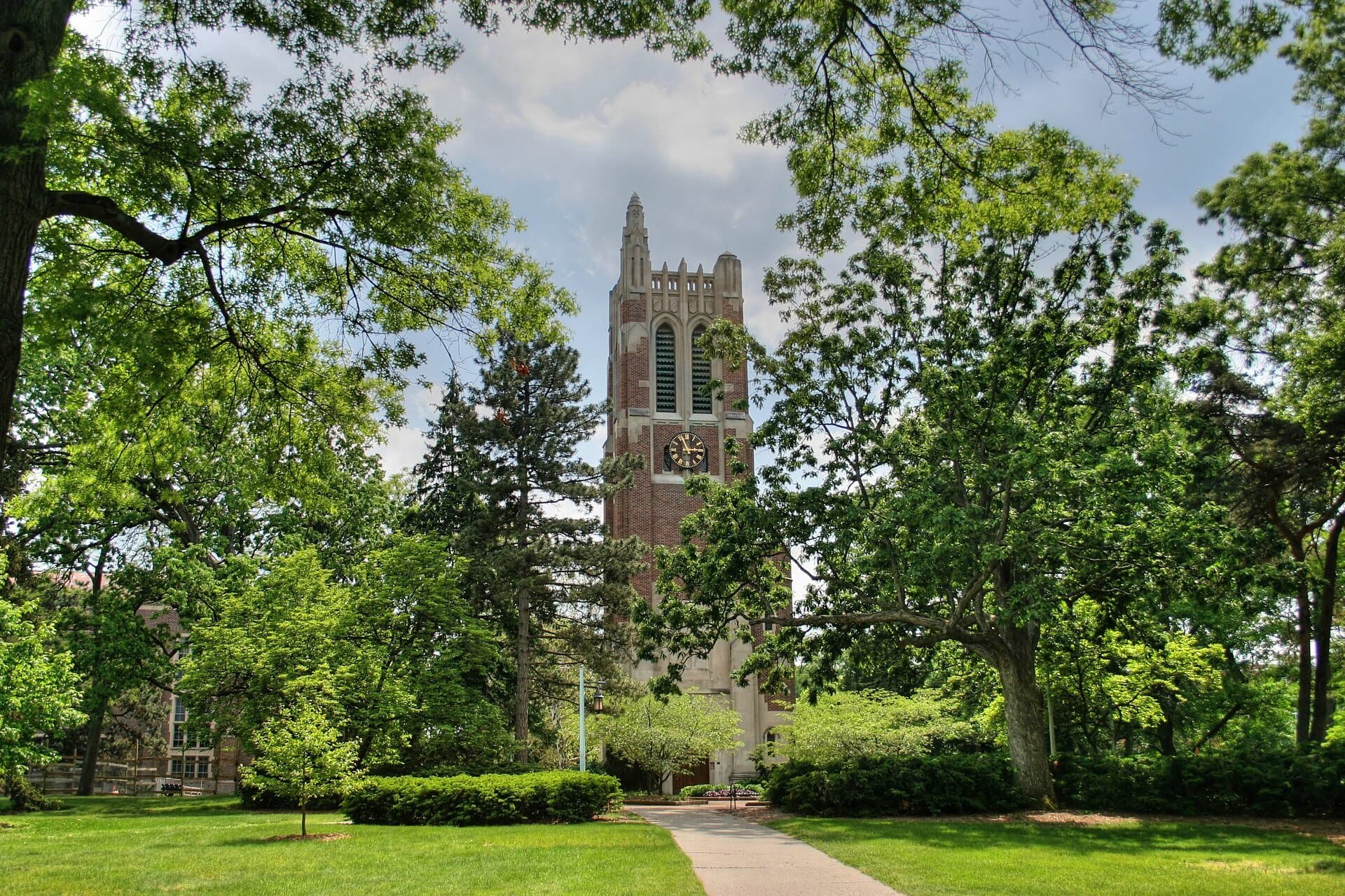 Michigan State's red-brick Beaumont Tower rising above tall green trees at the end of a lawn and walkway on a sunny day