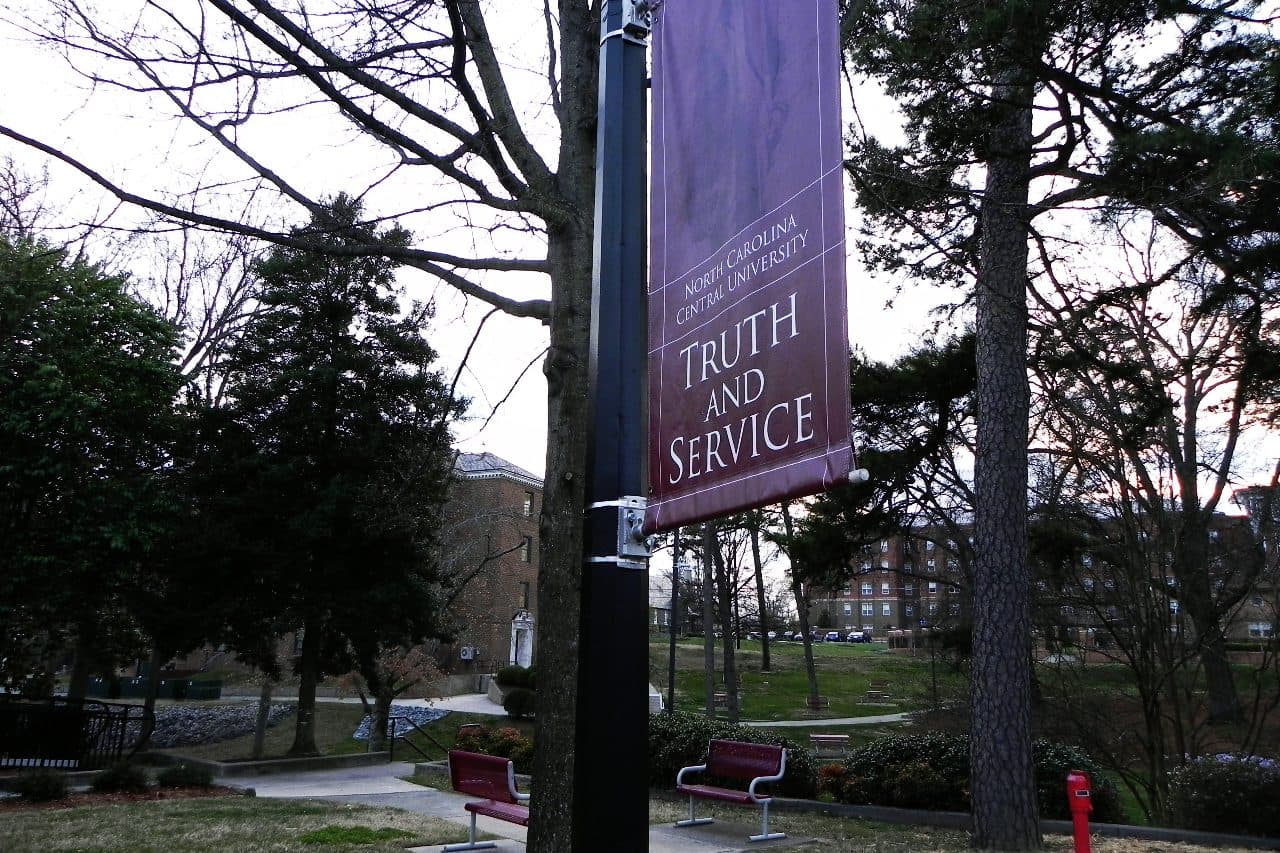 A tree-lined walkway on North Carolina Central University's campus with a maroon "Truth and Service" banner on a lamp post and brick buildings behind
