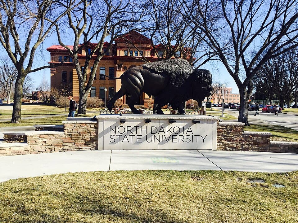 A bison statue on a stone sign reading North Dakota State University, with a red-brick building and bare trees behind it on a sunny day
