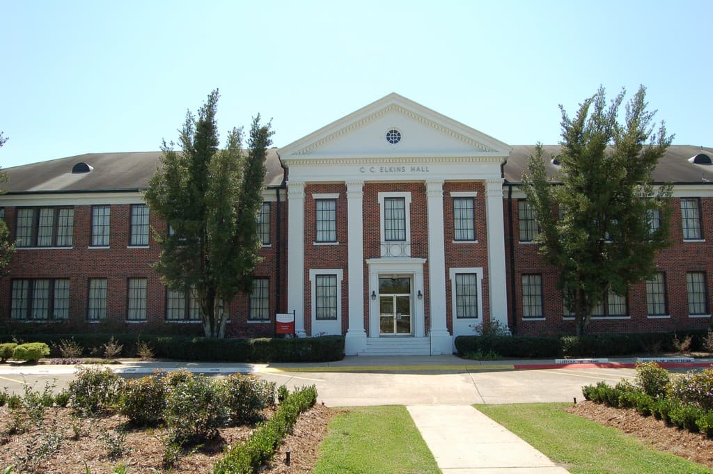 A two-story red-brick building with a white columned portico and triangular pediment reading C C Elkins Hall, framed by two tall trees, with a concrete walk and landscaped beds in front under a clear blue sky