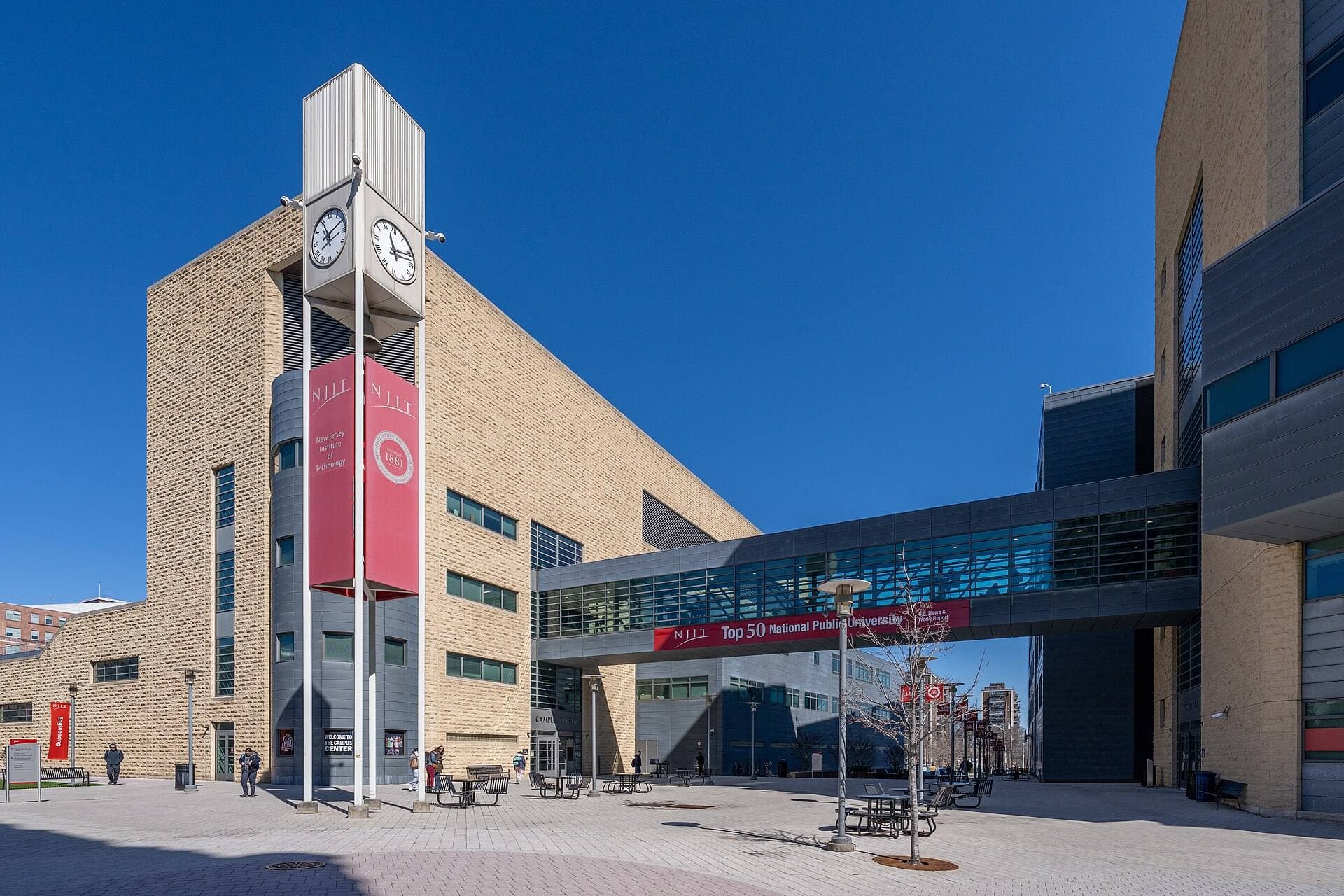 A tan brick clock tower with red banners beside a glass skybridge and a plaza on the NJIT campus in Newark