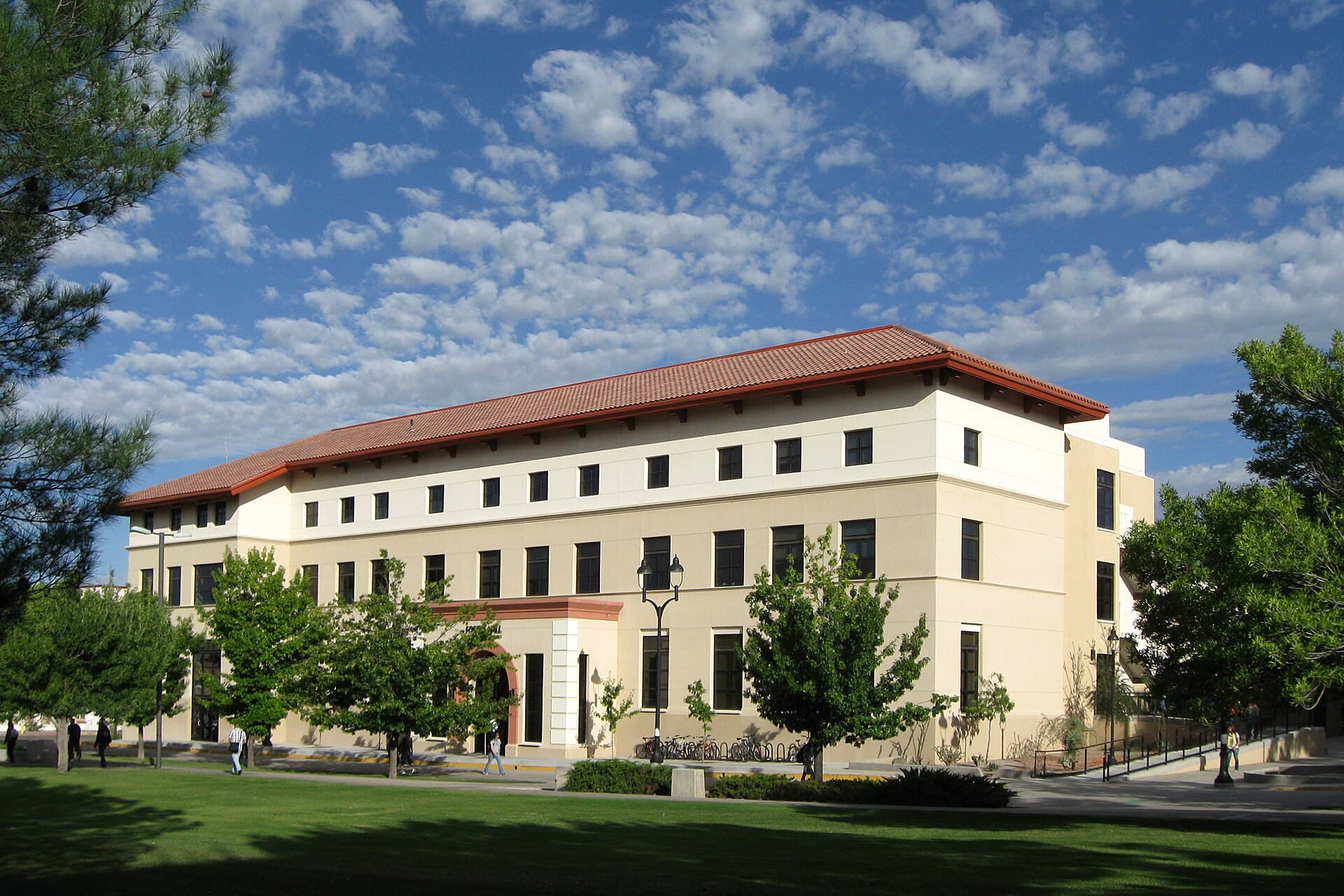 Foster Hall at New Mexico State University, a three-story cream stucco building with a red tile roof, behind a green lawn and trees under a partly cloudy sky
