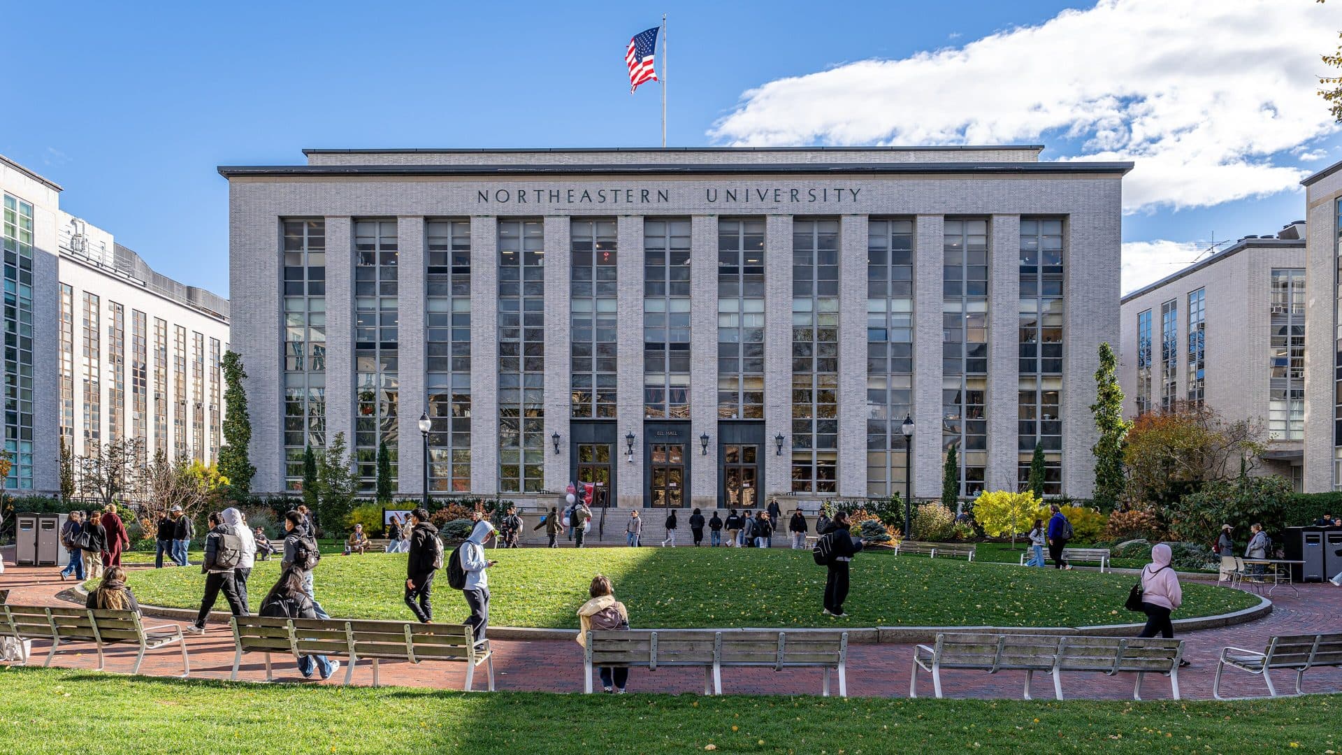 Ell Hall at Northeastern University, a pale stone building with tall windows and an American flag on the roof, behind a lawn where students walk on a sunny day