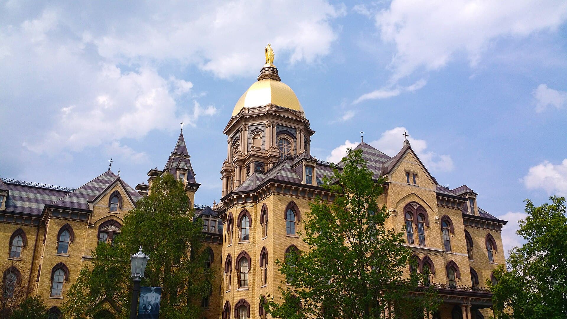 Notre Dame's cream-brick Main Building and its gold dome and statue rising above trees against a blue sky with white clouds