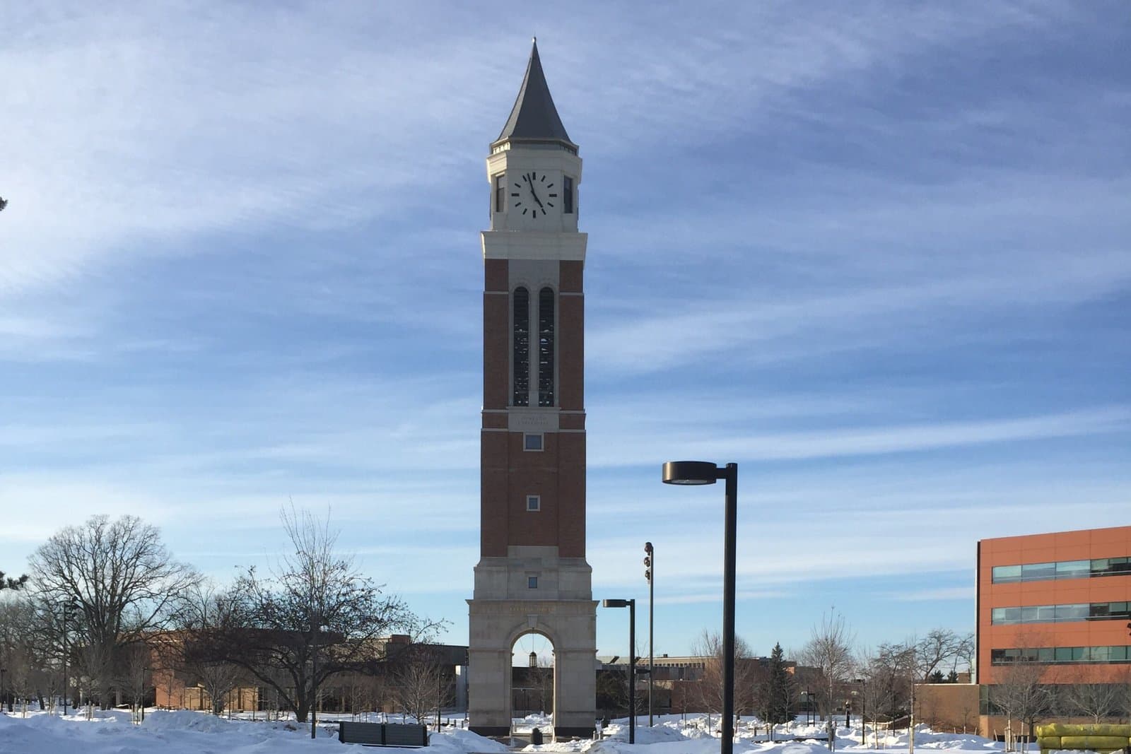 A tall red-brick carillon tower with a white stone arched base, a clock face and a pointed slate spire, standing at the end of a shoveled walkway between snowbanks under a blue sky streaked with thin clouds, with bare trees and campus buildings behind it