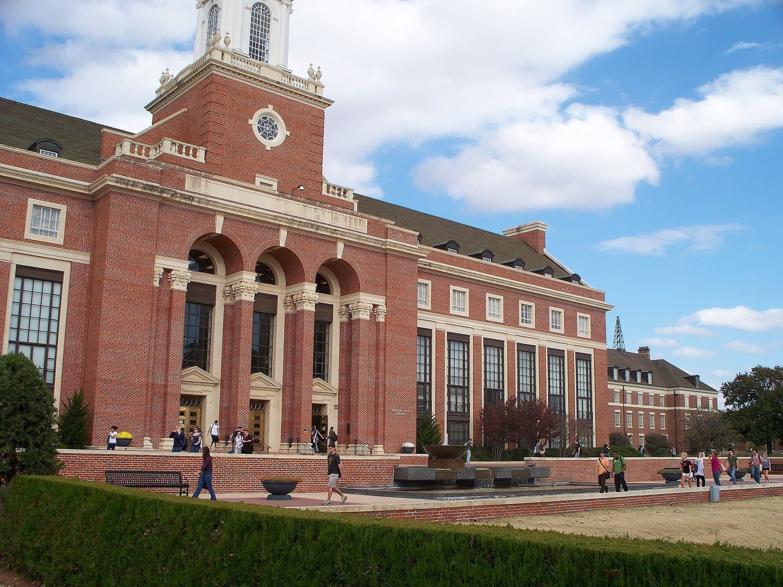 Students walking past the arched entrance and white clock tower of Edmon Low Library, a red-brick Georgian building, under a blue autumn sky at Oklahoma State University