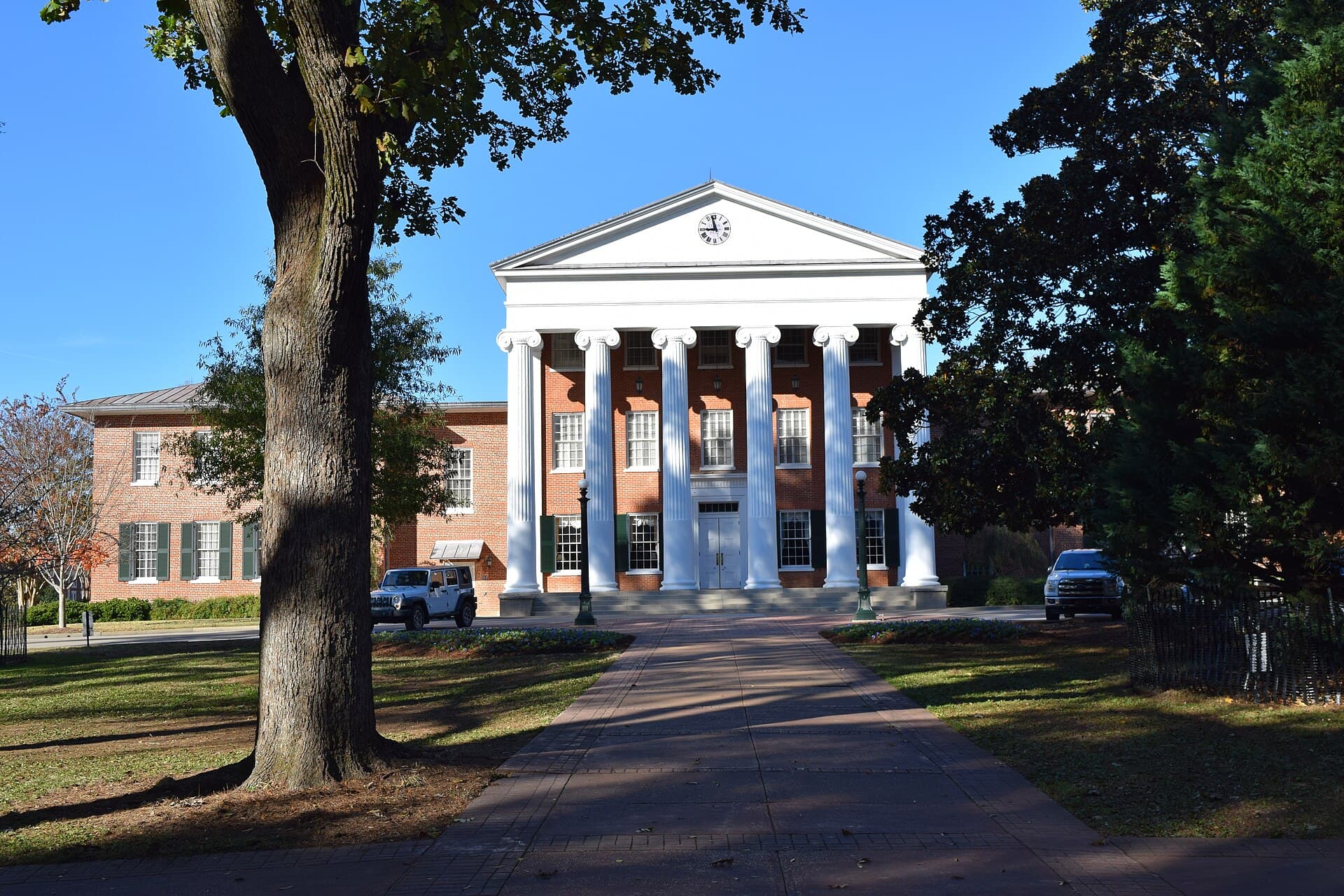 The Lyceum at Ole Miss, a red-brick building with six tall white columns and a clock in its pediment, at the end of a brick walk shaded by a large oak under a clear blue sky