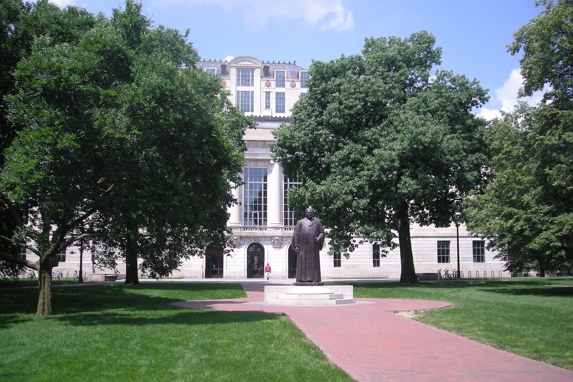 A brick path across the green Oval at Ohio State, lined with large shade trees, leading to a statue and the columned front of Thompson Library