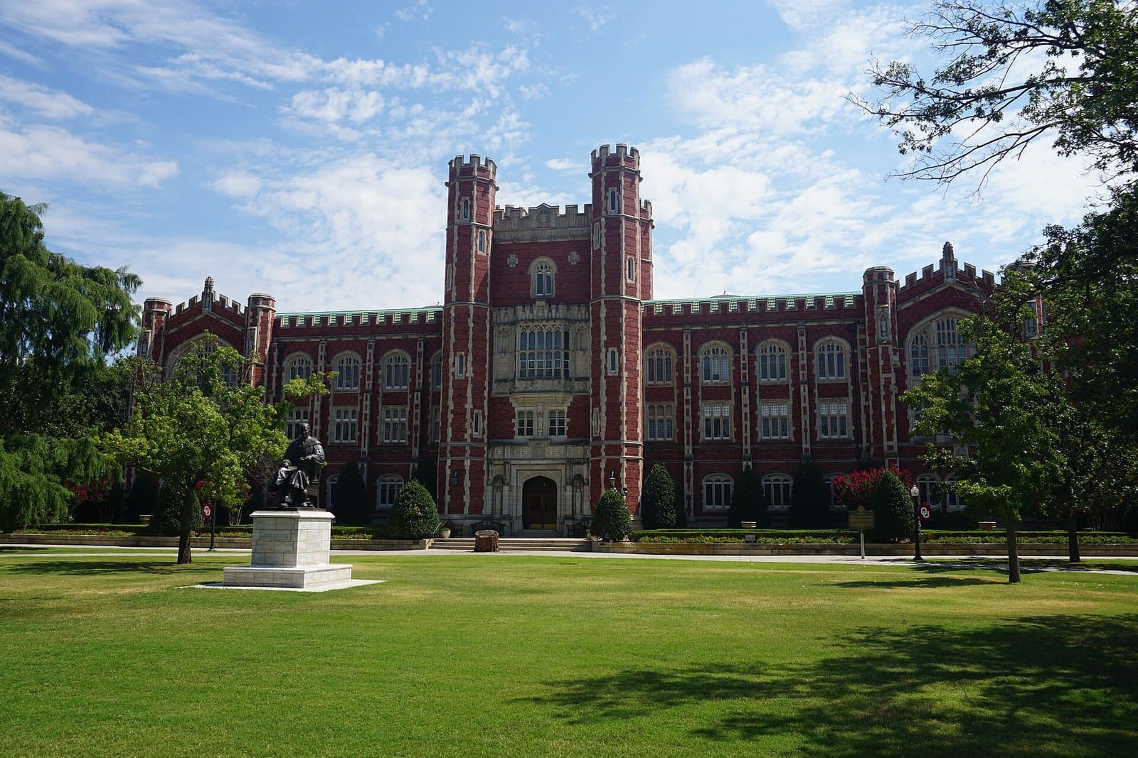 Evans Hall, a red-brick Gothic building with crenellated twin towers, behind a wide green lawn and a seated bronze statue at the University of Oklahoma