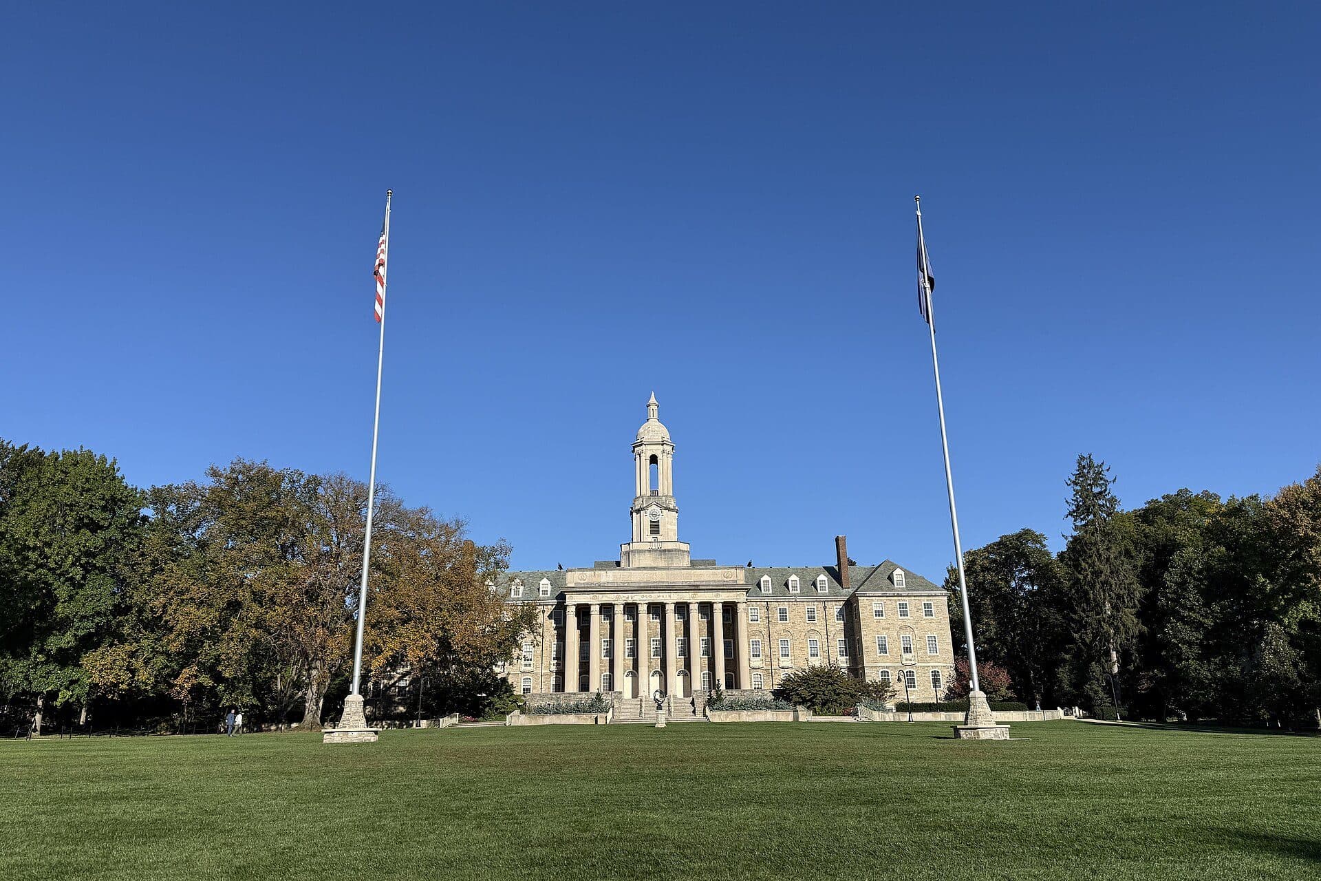 Old Main at Penn State, a stone building with a white bell tower, seen across the Old Main Lawn with flagpoles under a blue sky