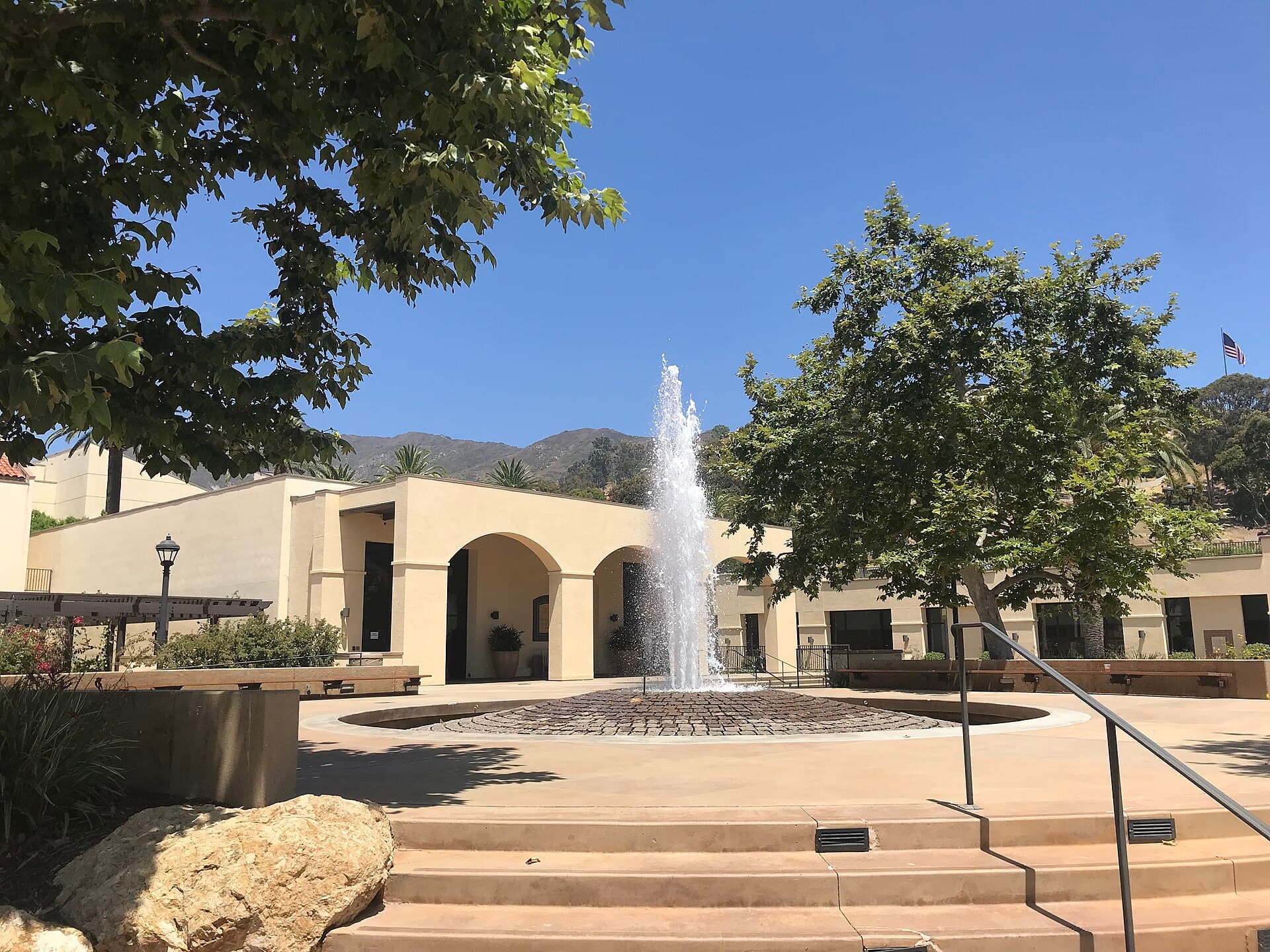 A tall fountain jet rising from a round brick-paved basin in a sunny plaza, with cream stucco buildings with arched entries behind it, shade trees on both sides, wide steps in the foreground and the Santa Monica Mountains under a clear blue sky