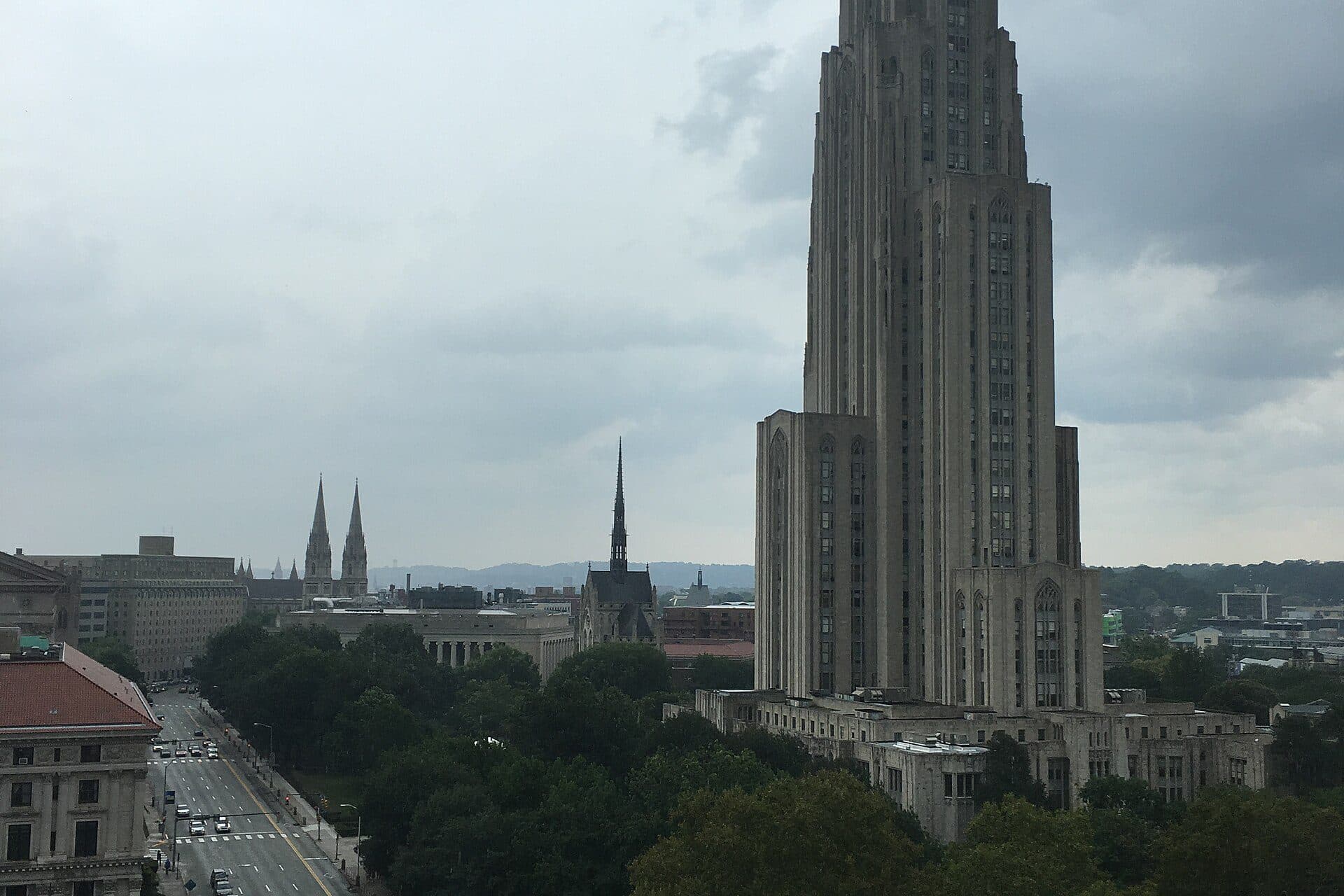 The Gothic Revival Cathedral of Learning tower rising above Oakland under a cloudy sky, with church spires and city streets around it
