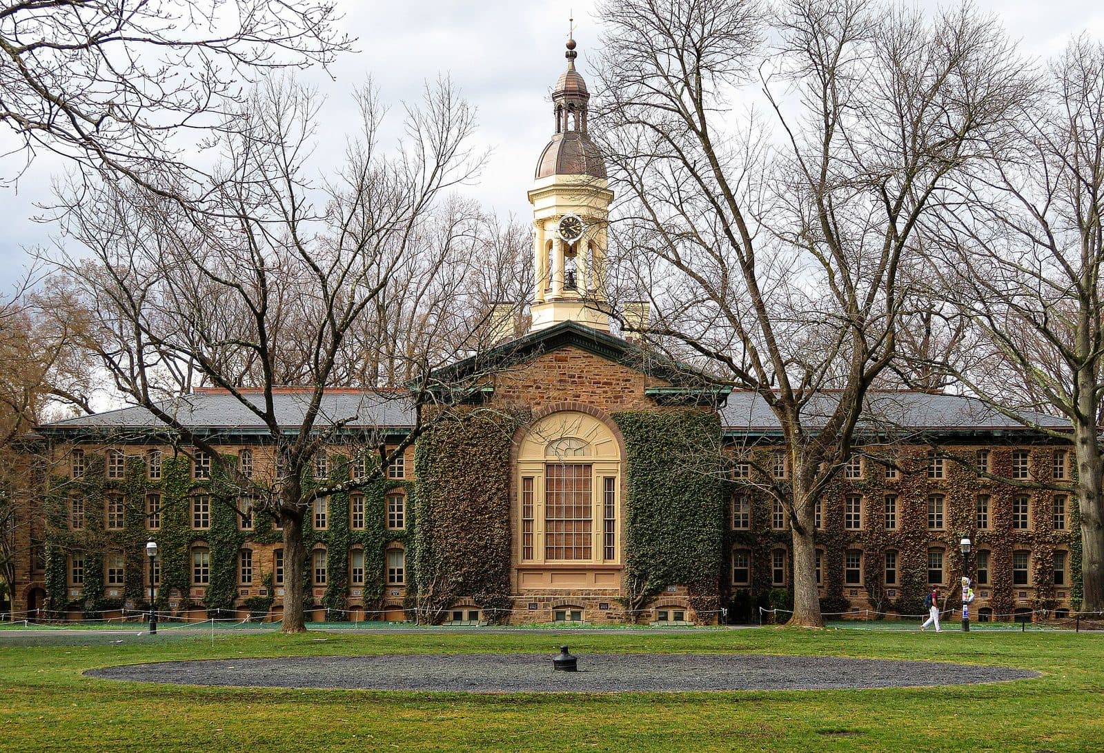 Ivy-covered Nassau Hall, a long stone building with a white cupola and clock, seen across a green lawn and bare early-spring trees at Princeton University, under a cloudy sky