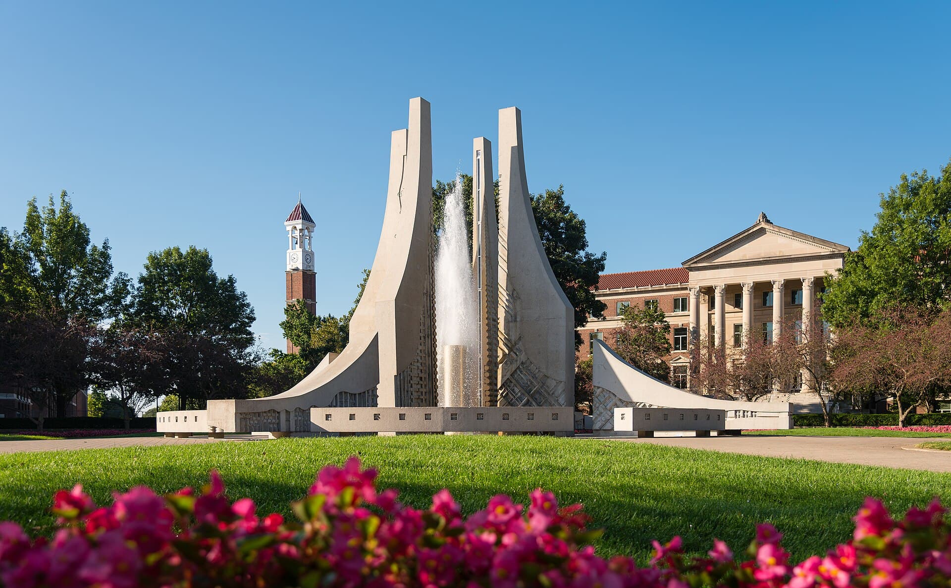 Purdue's Engineering Fountain, tall curving pale stone fins with a jet of water, behind a lawn and pink flowers, with the red-brick Purdue Bell Tower on the left and columned Hovde Hall on the right under a clear blue sky
