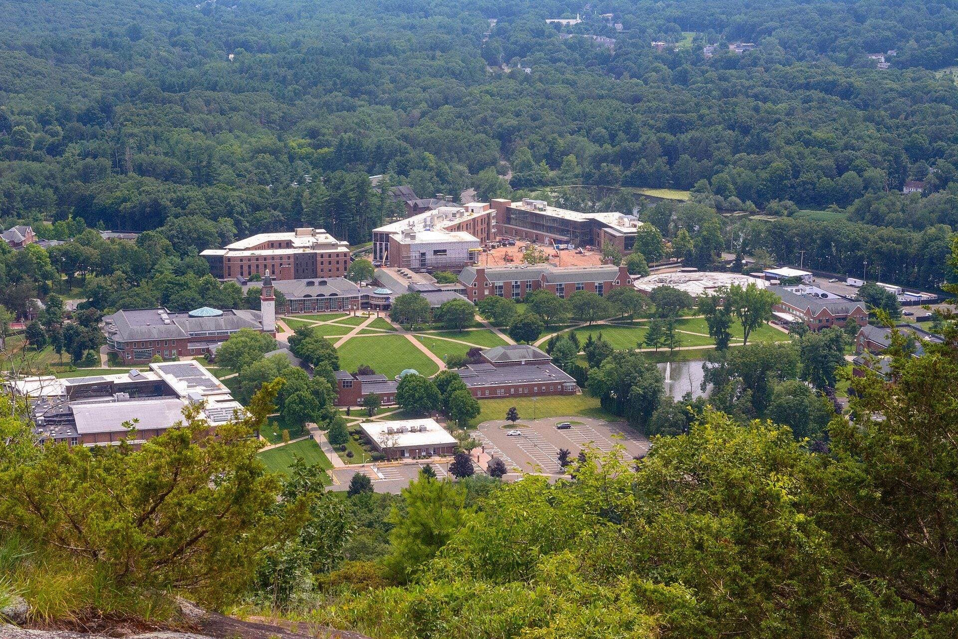 View from a wooded ridge at Sleeping Giant State Park down to Quinnipiac's Mount Carmel Campus, with brick buildings and green lawns surrounded by forested hills