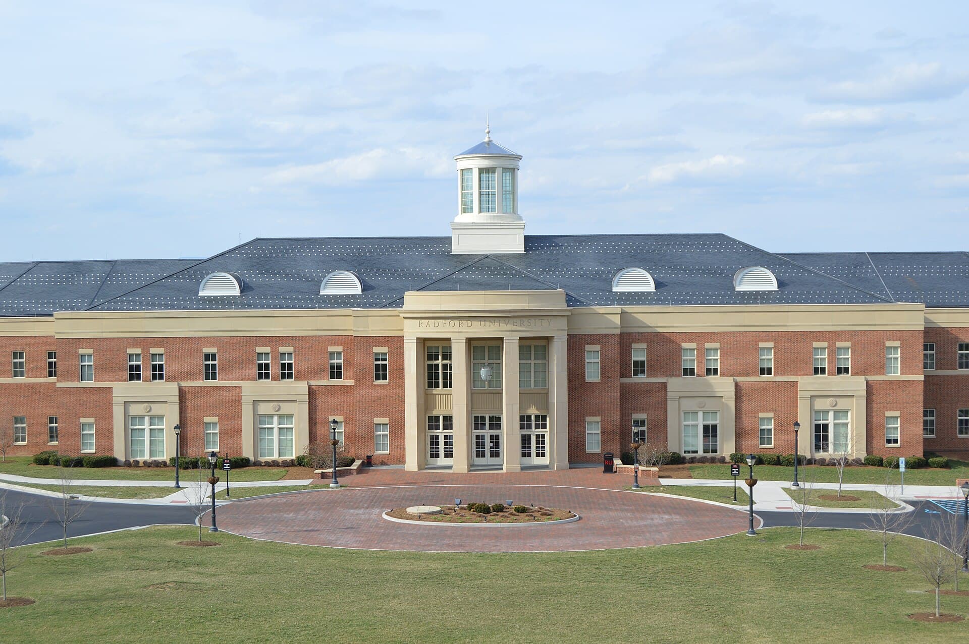 A long red-brick academic building with cream stone trim, a columned center entrance with RADFORD UNIVERSITY carved above it, a white cupola on a gray slate roof, and a brick circle drive and lawn in front under a pale blue sky