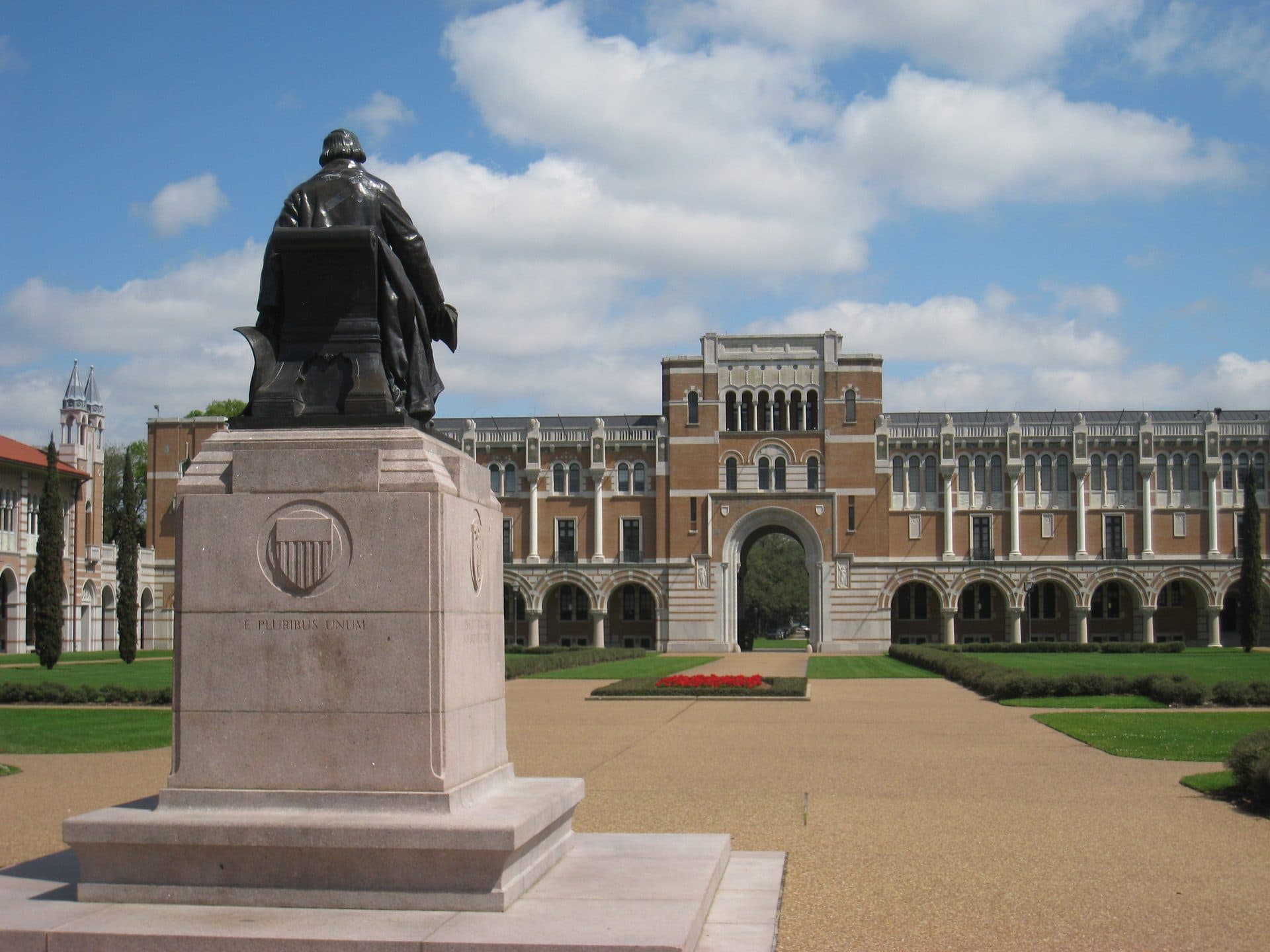 The bronze William Marsh Rice statue seen from behind, facing Lovett Hall's long arched brick-and-stone facade across a lawn