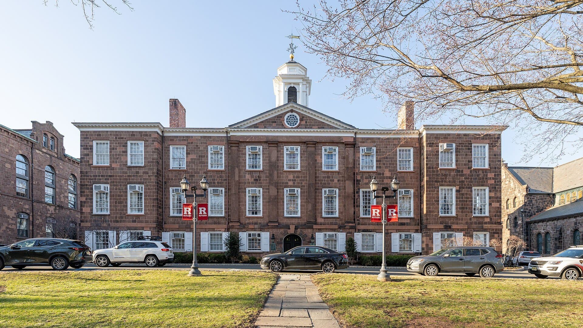 Old Queens at Rutgers, a three-story brownstone building with rows of white-framed windows and a white cupola, with red Rutgers banners on lampposts and a lawn and walkway in front under a clear sky
