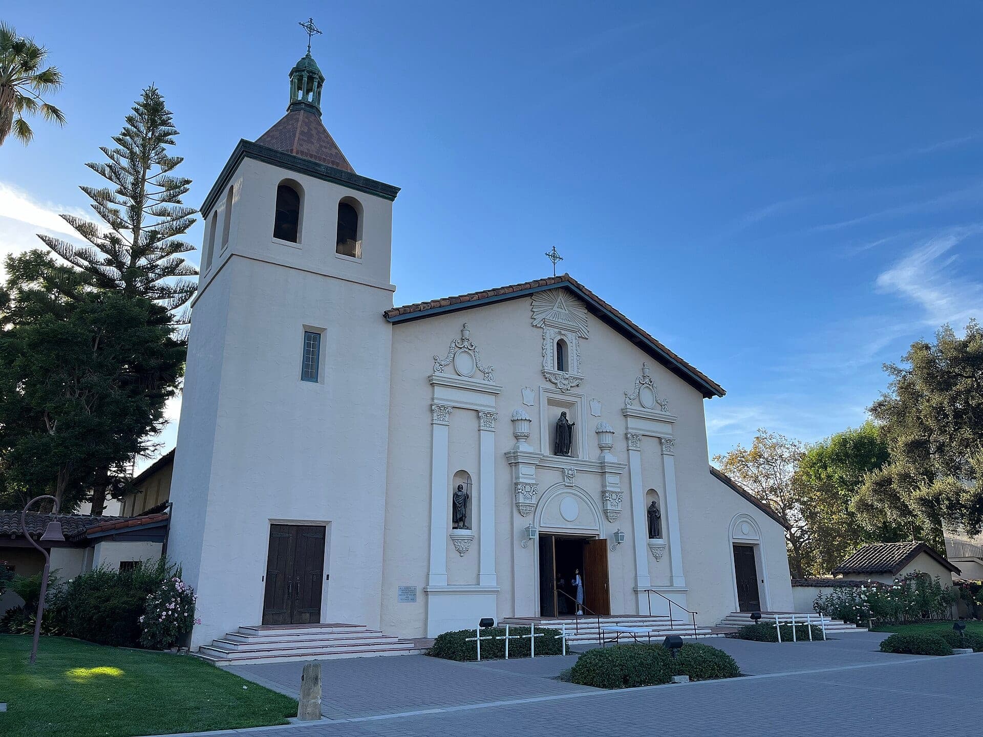 The white facade and bell tower of Mission Santa Clara de Asís in Santa Clara, California, framed by trees under a blue sky