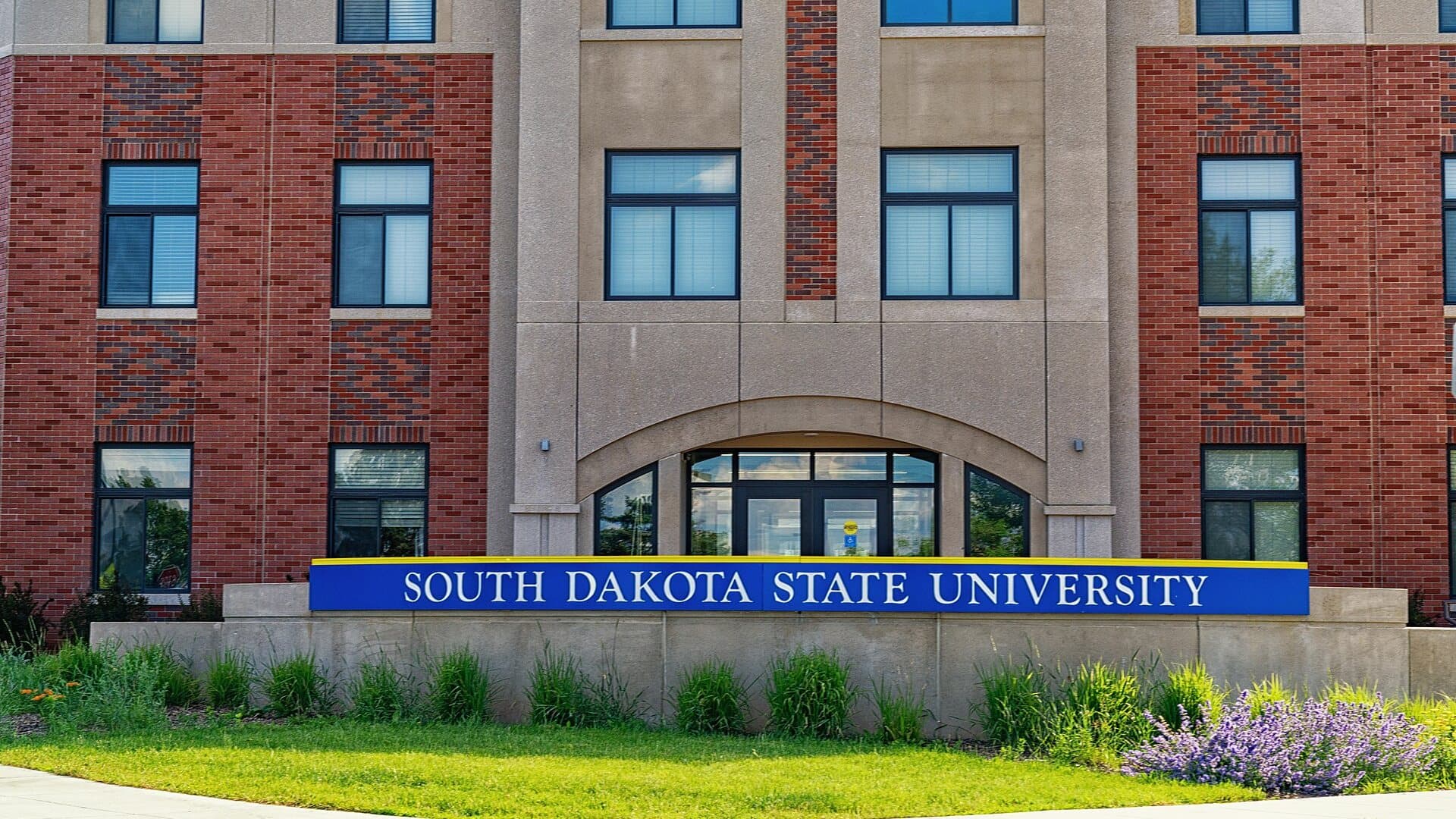 A long royal-blue sign with gold trim reading South Dakota State University on a low stone wall, in front of a red-brick and tan stone building with an arched glass entrance, with green lawn, ornamental grasses and purple flowers under summer sun