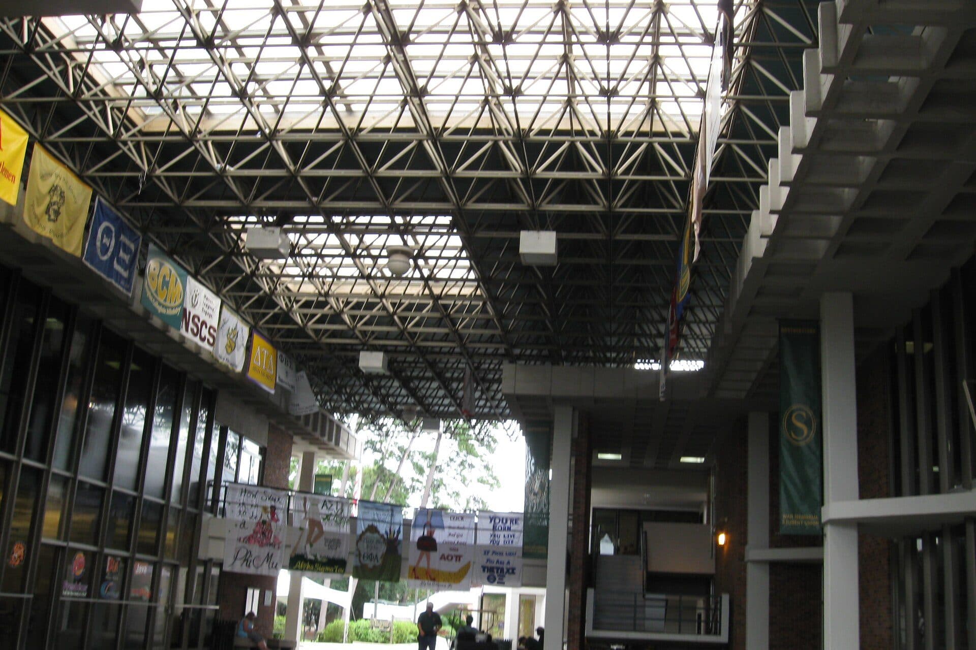 A covered breezeway with a steel-truss skylight roof running alongside the Southeastern Louisiana University Student Union, hung with colorful banners