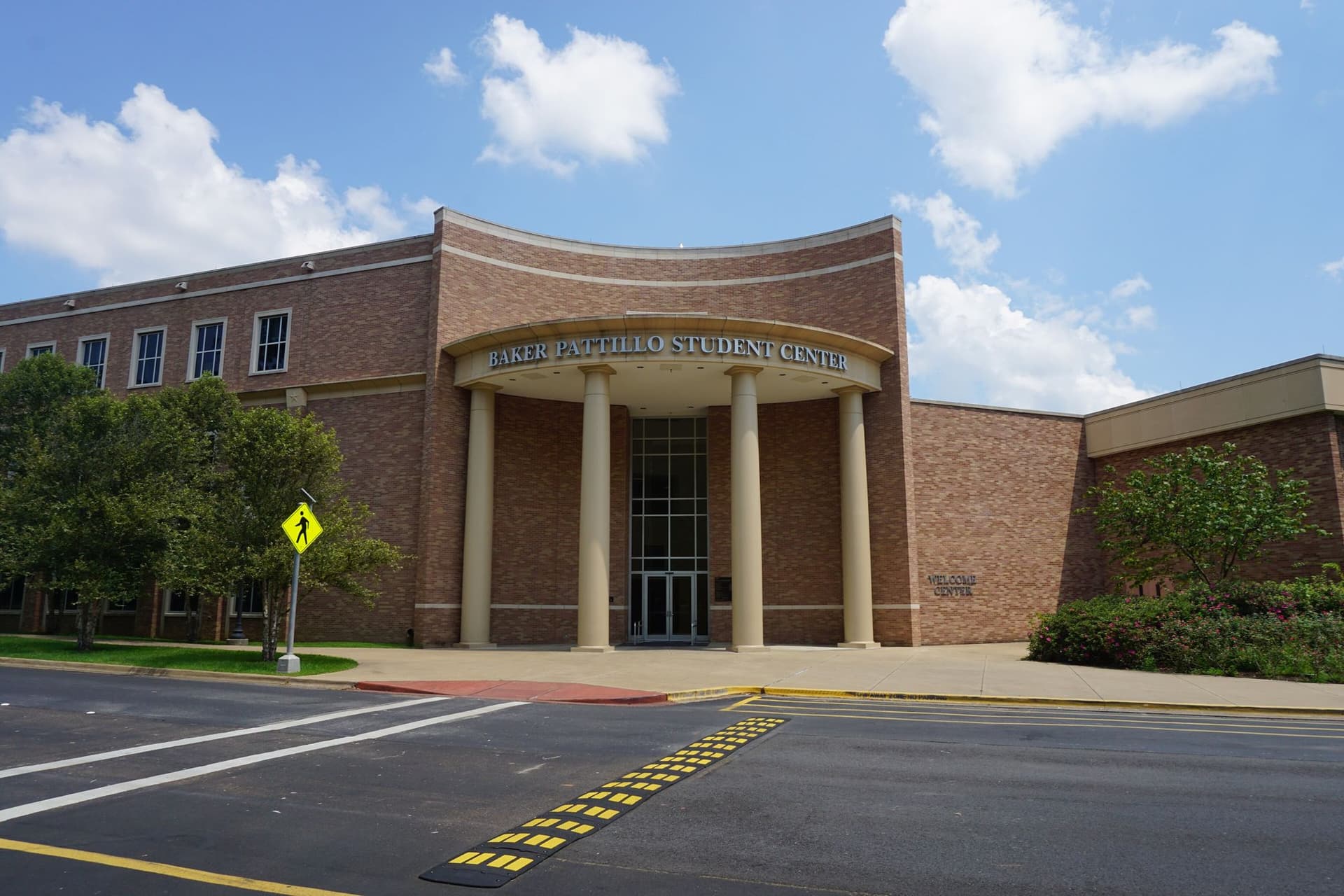 A reddish-brown brick building with a curved, columned entrance under a sign reading Baker Pattillo Student Center, with a crosswalk and speed bump in the road in front, green trees and shrubs at either side and white clouds in a blue sky