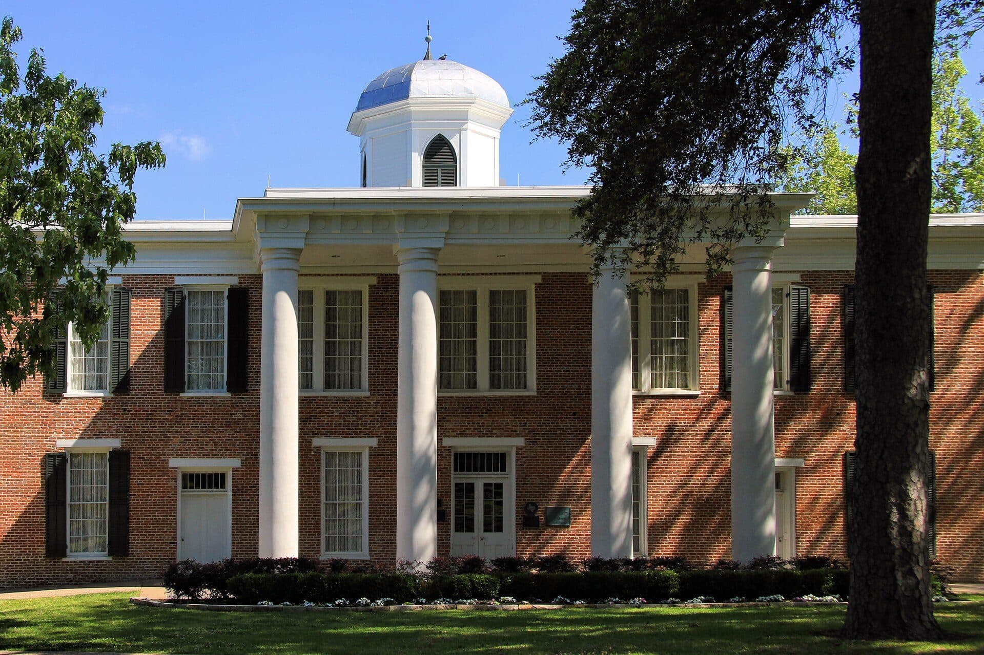 Sam Houston State's red-brick Austin Hall with tall white columns, black shutters and a white domed cupola, behind a green lawn