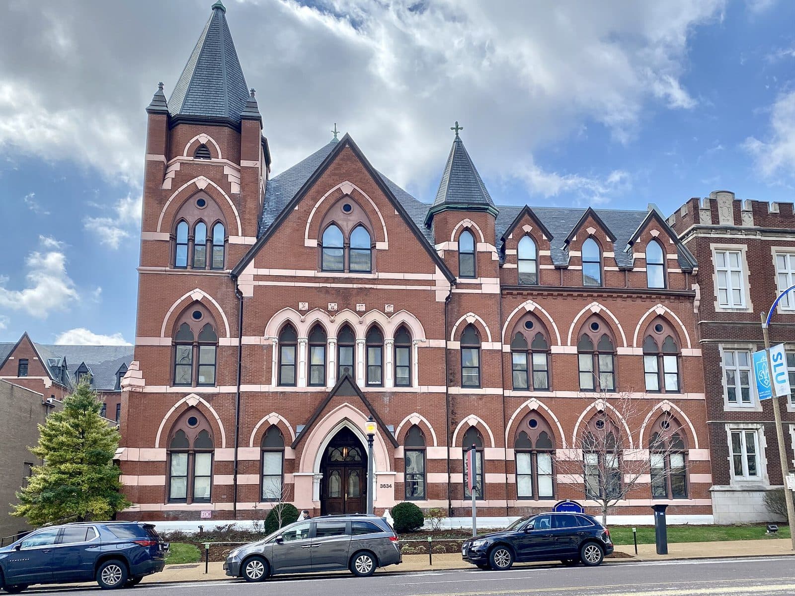 Verhaegen Hall, a red-brick Gothic Revival building with pointed-arch windows, a tall slate-roofed spire and a smaller cross-topped turret on Lindell Boulevard at Saint Louis University, with cars parked along the street and a blue SLU banner on a lamppost under a partly cloudy sky