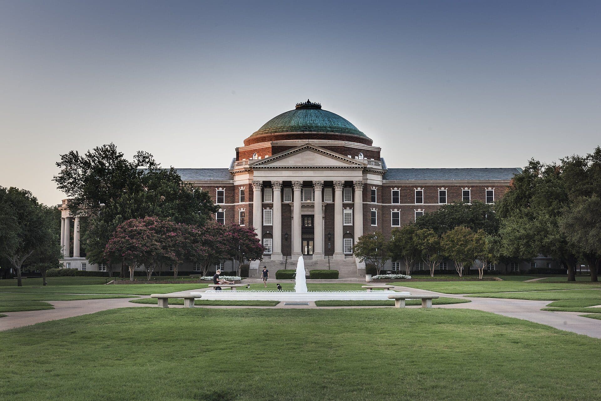 Dallas Hall's red-brick facade, white columns and green copper dome at dusk, behind a fountain and lawn on the SMU campus