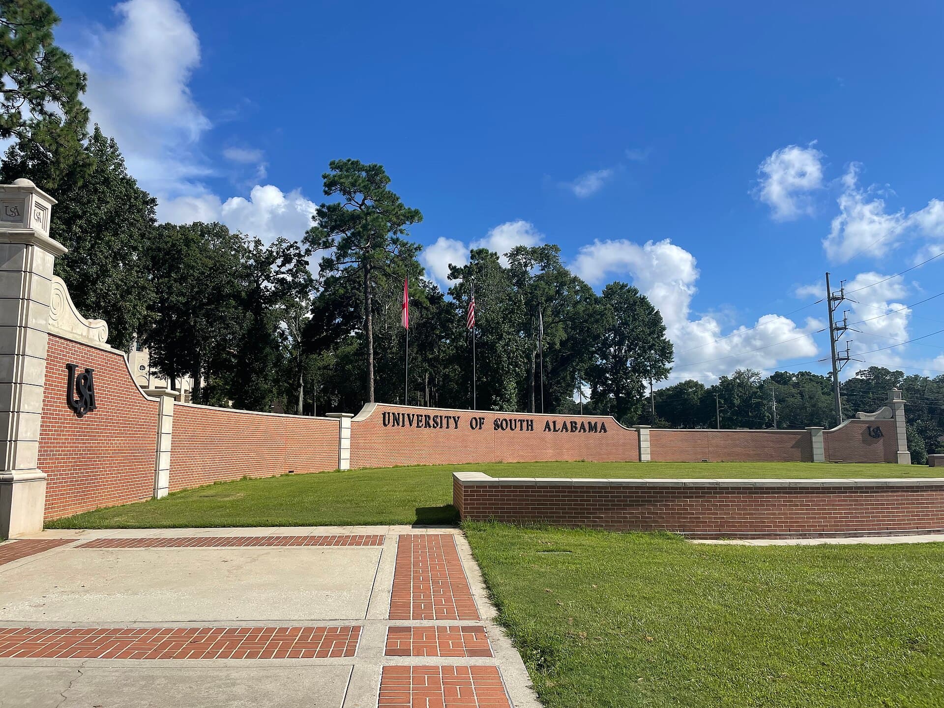 A long red-brick wall with stone trim reading UNIVERSITY OF SOUTH ALABAMA, with USA monograms, a brick-and-concrete walkway and green lawn in front and tall pines and flags behind under a blue sky with white clouds