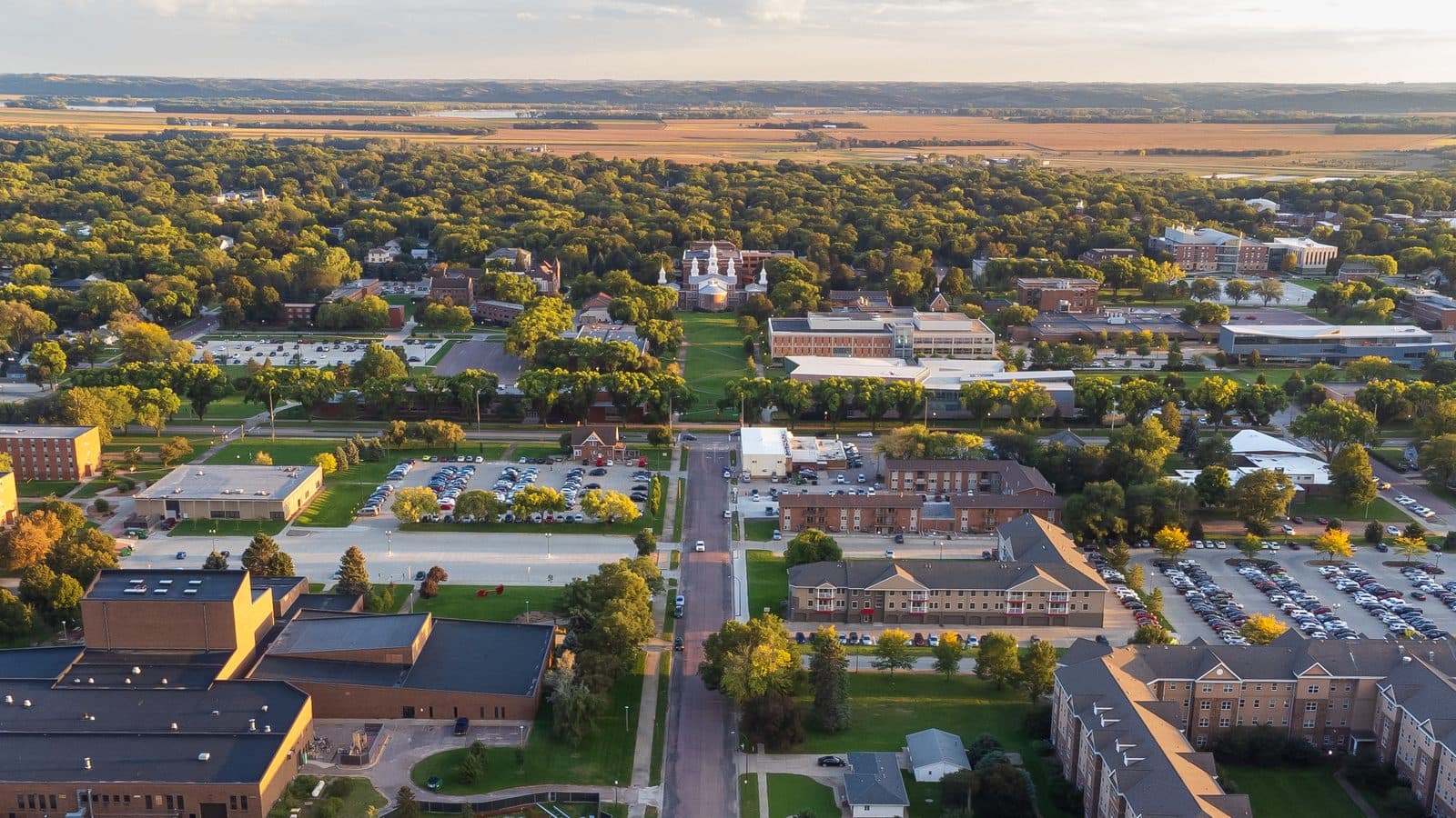 An aerial view at golden hour over a campus of red-brick buildings, parking lots and tree-lined streets, with a white-towered historic hall among green trees and farm fields stretching to the horizon