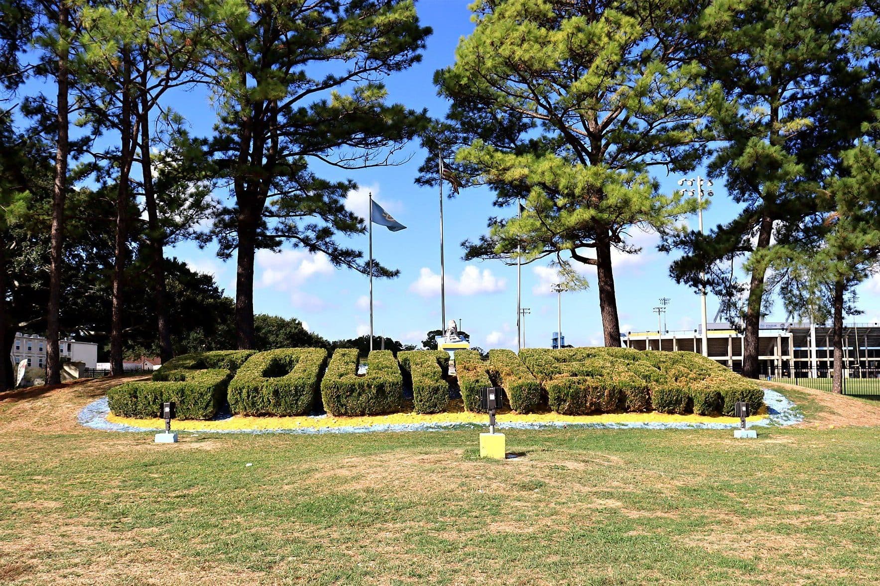 Trimmed hedges spelling out SU on a lawn at the Southern University entrance, framed by tall pine trees under a blue sky