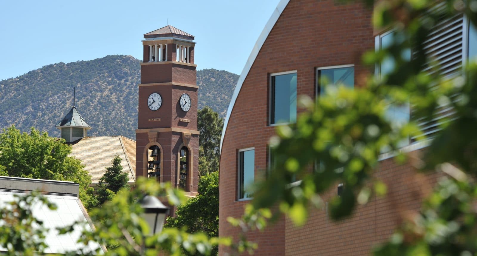 The red-brick Carter Carillon bell tower with its clock faces, framed by green leaves and a curved brick building, with juniper-covered hills behind at Southern Utah University