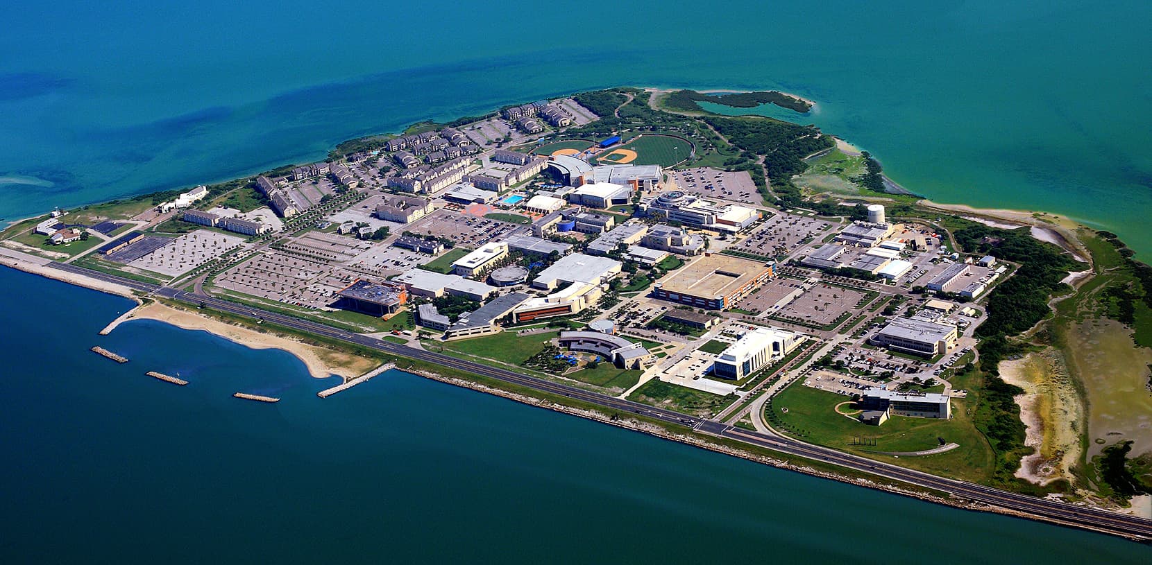 Aerial view of a campus of white and tan buildings, parking lots, athletic fields and residence halls on a green peninsula, with turquoise bay water on three sides and a shoreline road and small beach along the front
