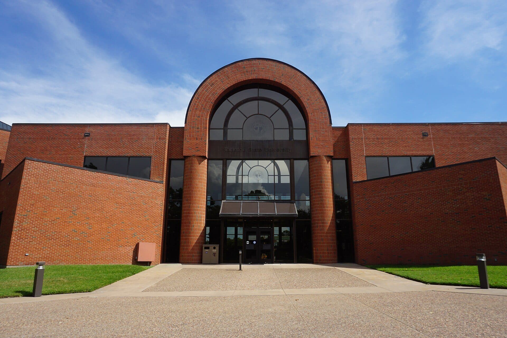 A red-brick building with a tall central arched entrance framed by two round brick columns and a large half-round window, flanked by low brick wings, with a wide concrete walkway and green lawns in front under a blue sky with thin clouds
