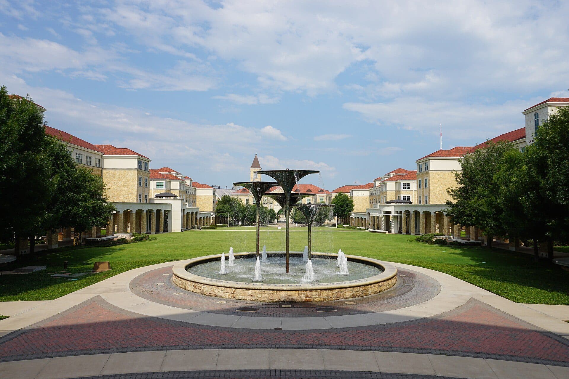 A broad green lawn at TCU framed by matching tan-brick buildings with red tile roofs and arched arcades on both sides