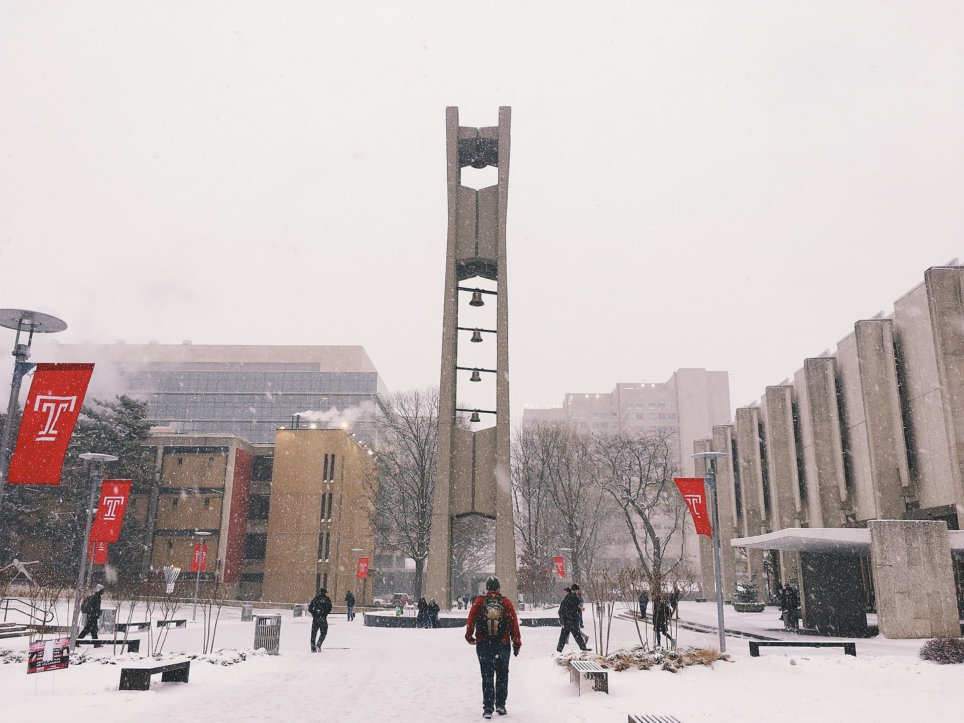 Temple University's tall concrete Bell Tower on a snowy campus plaza, with red Temple T banners on light poles and students walking through the snow