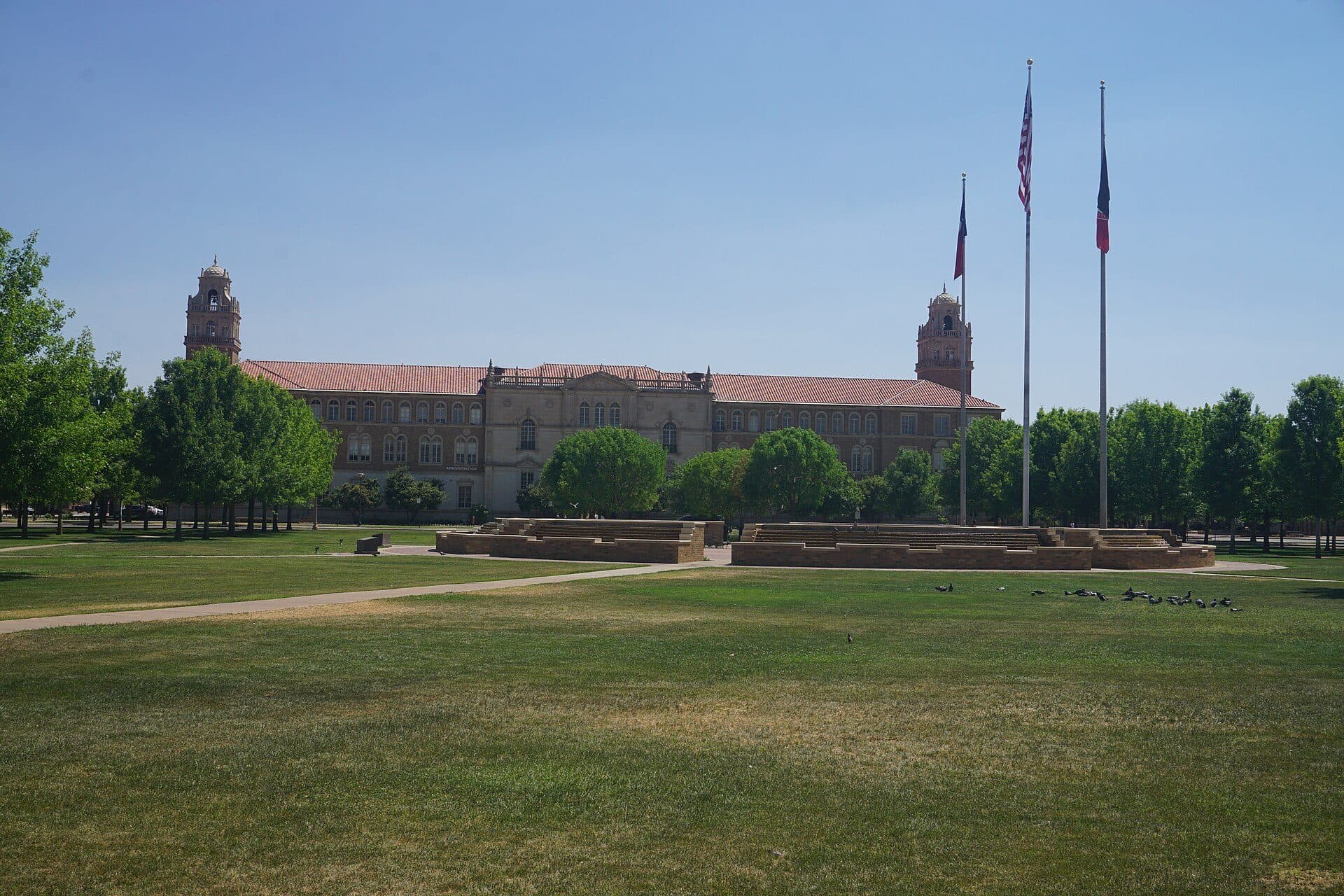 Texas Tech's Spanish Renaissance-style Administration Building, with twin bell towers and a red tile roof, seen across the lawn of Memorial Circle with flagpoles and trees