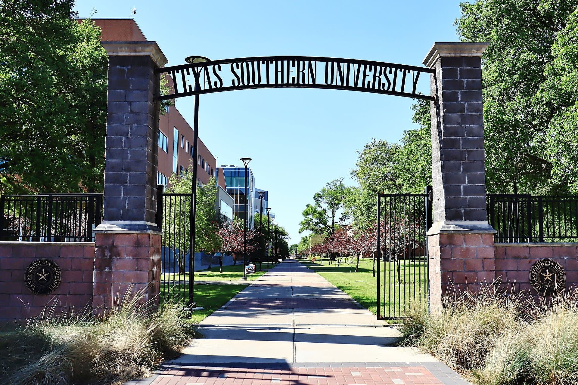 A black iron arch reading Texas Southern University between brick and stone pillars, with a brick walkway leading into a green, tree-lined campus