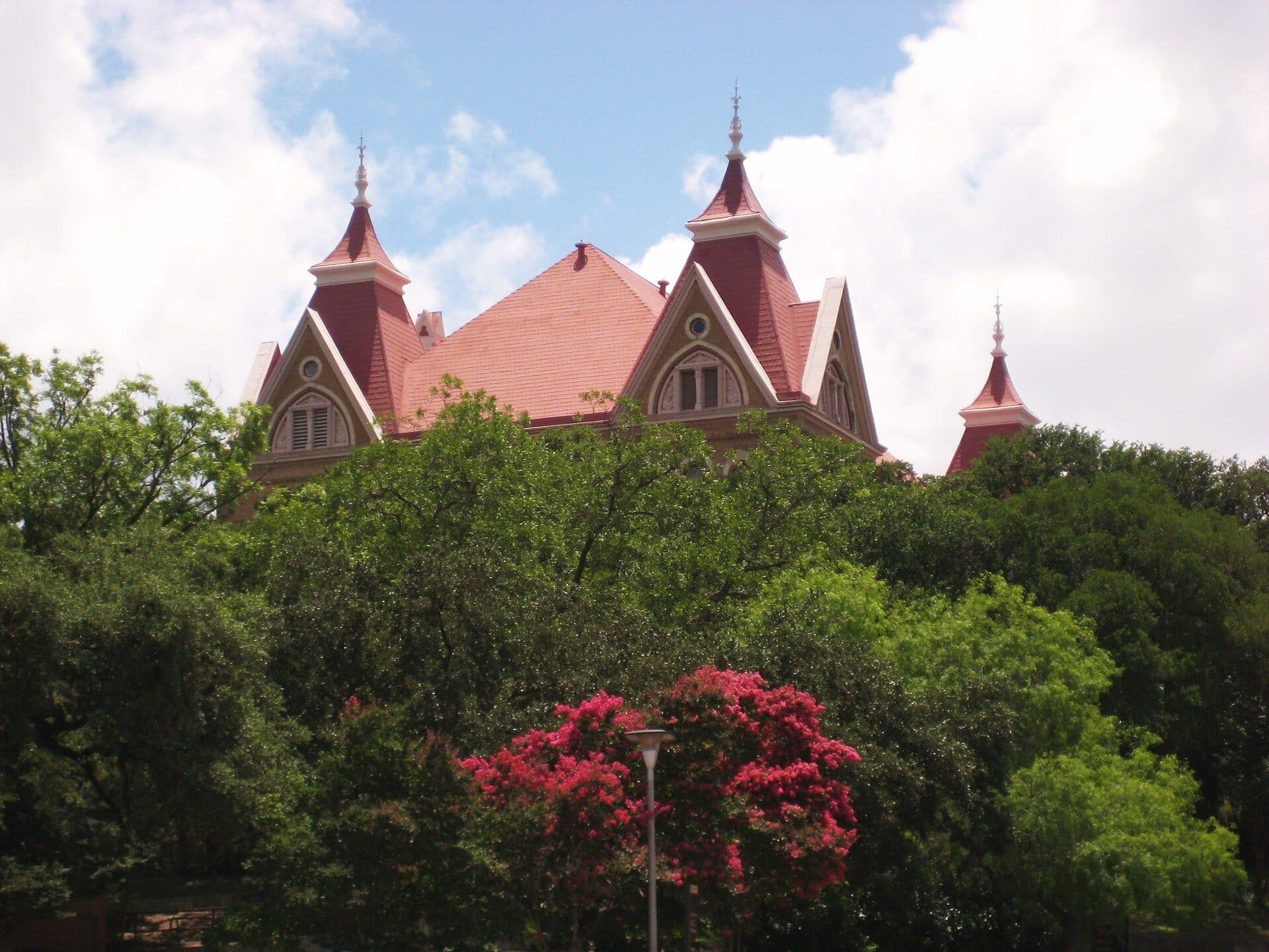 Old Main at Texas State University, a red-brick Victorian building with a red roof and pointed turrets, rising above green trees and pink crape myrtle blossoms