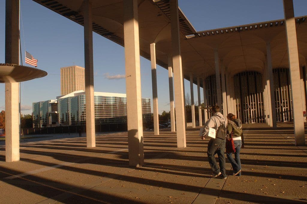 Two students walk beneath the tall white columns and canopy of the Academic Podium on UAlbany's Uptown Campus in late-afternoon light