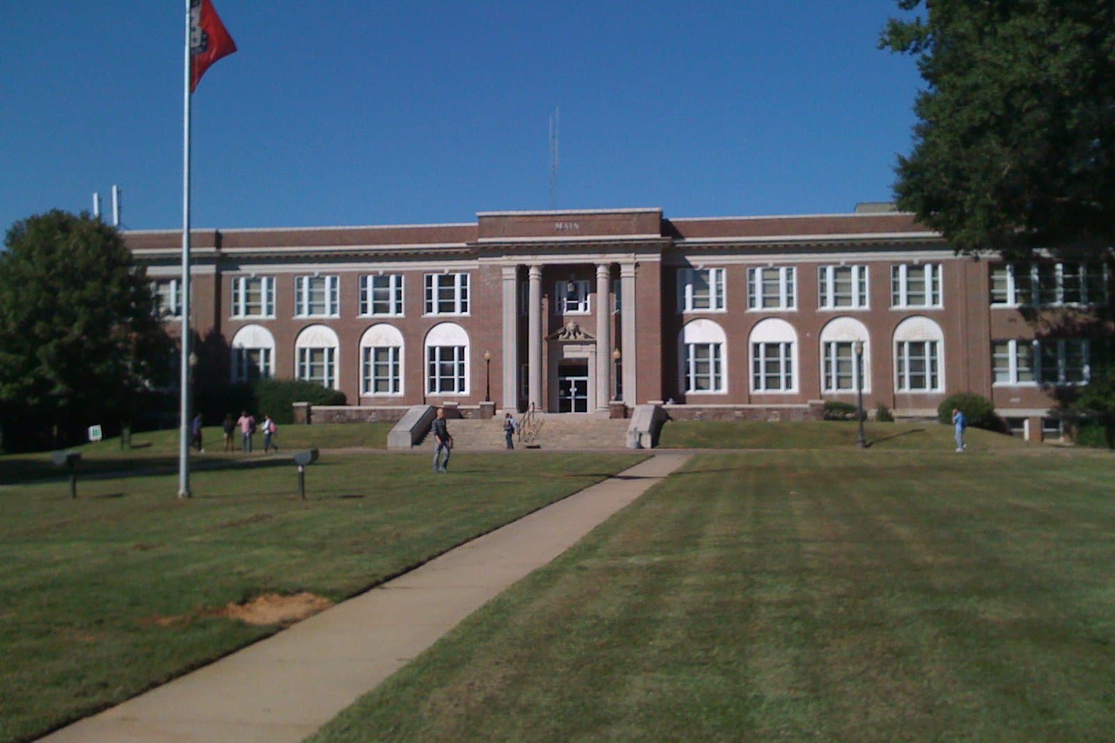 A long two-story red-brick building with tall arched windows, a columned central entrance and a parapet reading Main, behind a wide striped lawn crossed by a concrete walkway, with a flagpole, a few students walking and leafy trees under a clear blue sky
