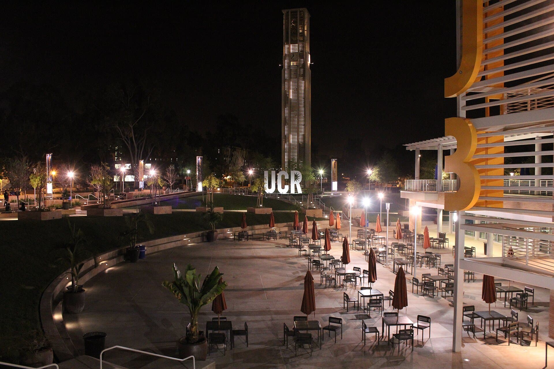 UC Riverside's Bell Tower lit up at night, with glowing UCR letters and a patio of tables and umbrellas in front