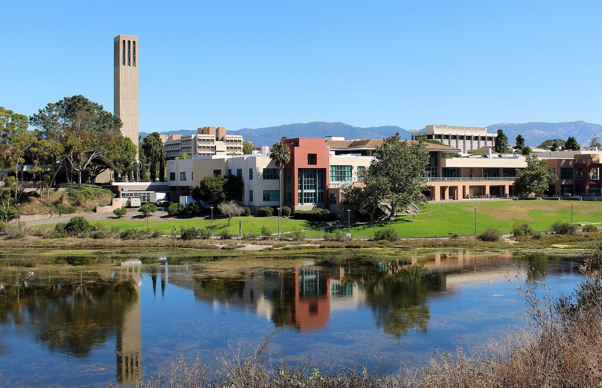 Storke Tower and the University Center at UC Santa Barbara reflected in the campus lagoon, with mountains in the distance under a blue sky