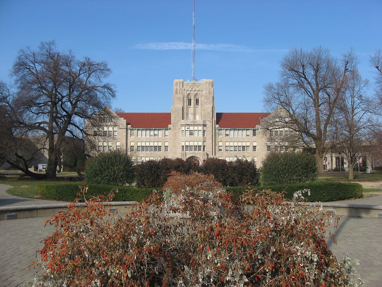 A pale limestone collegiate Gothic building with a central square tower, a tall antenna mast and red tile roofs, seen across a brick plaza with a round hedge-lined planter of red-berried shrubs in front, flanked by bare winter trees under a clear blue sky