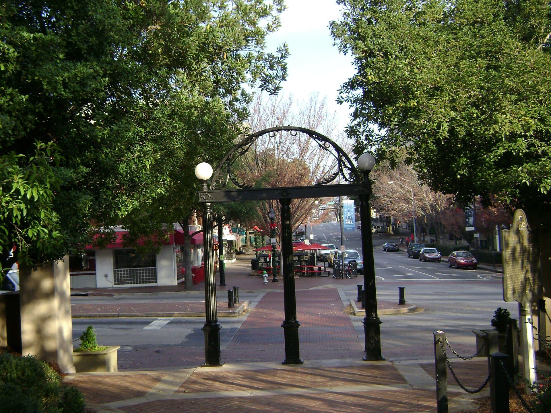 The black cast-iron Arch at the University of Georgia, with two lamp globes, on a brick walkway framed by green trees, looking out toward Broad Street and downtown Athens