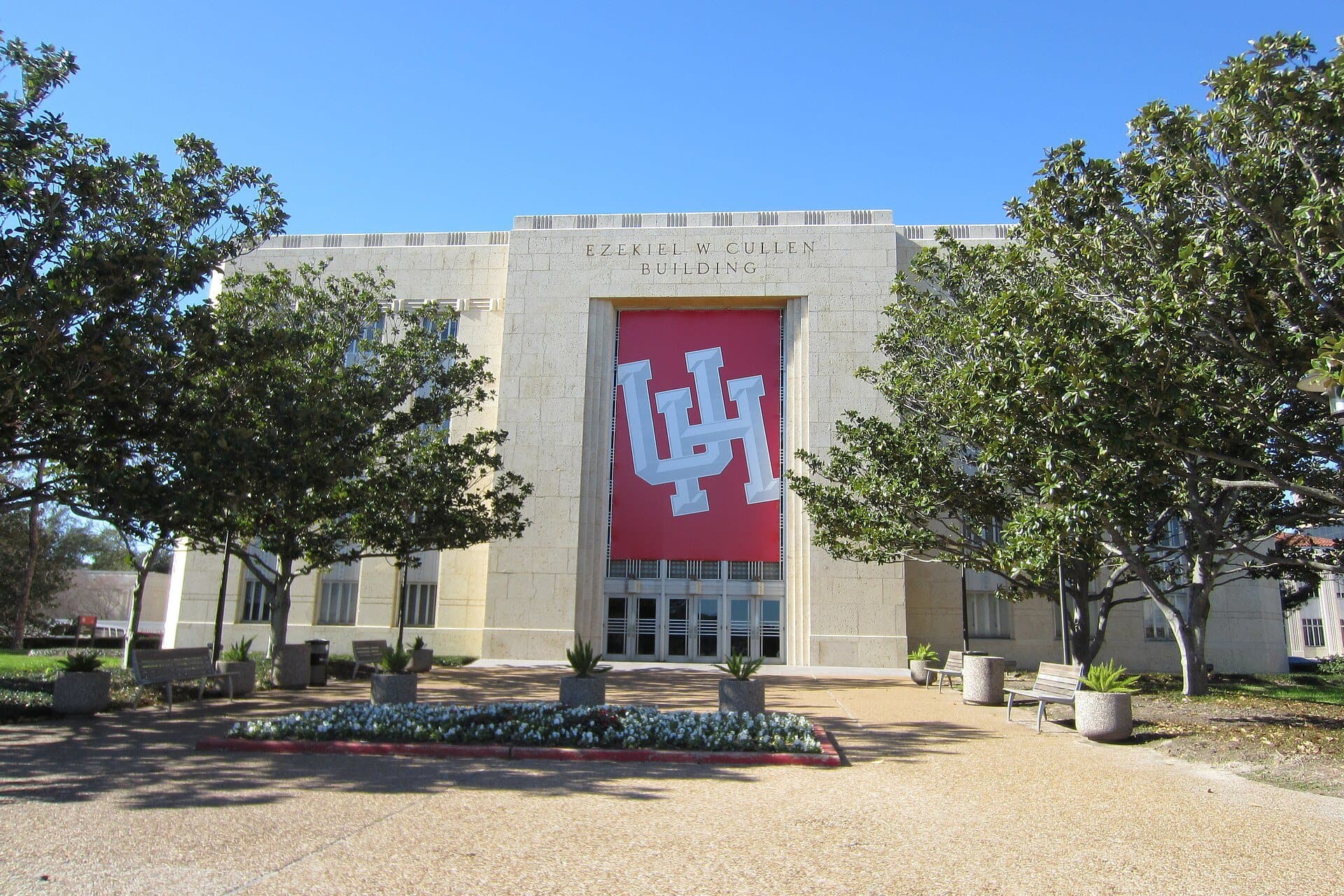 The pale limestone Ezekiel W. Cullen Building with a tall red UH banner over its entrance, framed by live oaks and a flower bed under a clear blue sky at the University of Houston